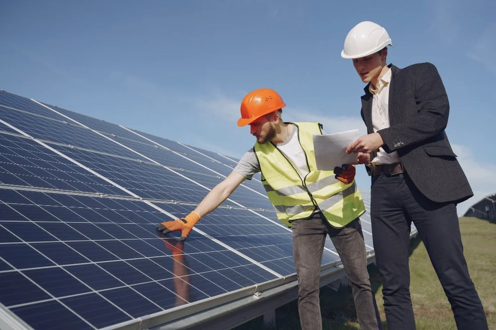 Two solar engineers, one in a safety vest and orange helmet, another in a suit and white helmet, inspecting solar panels outdoors.