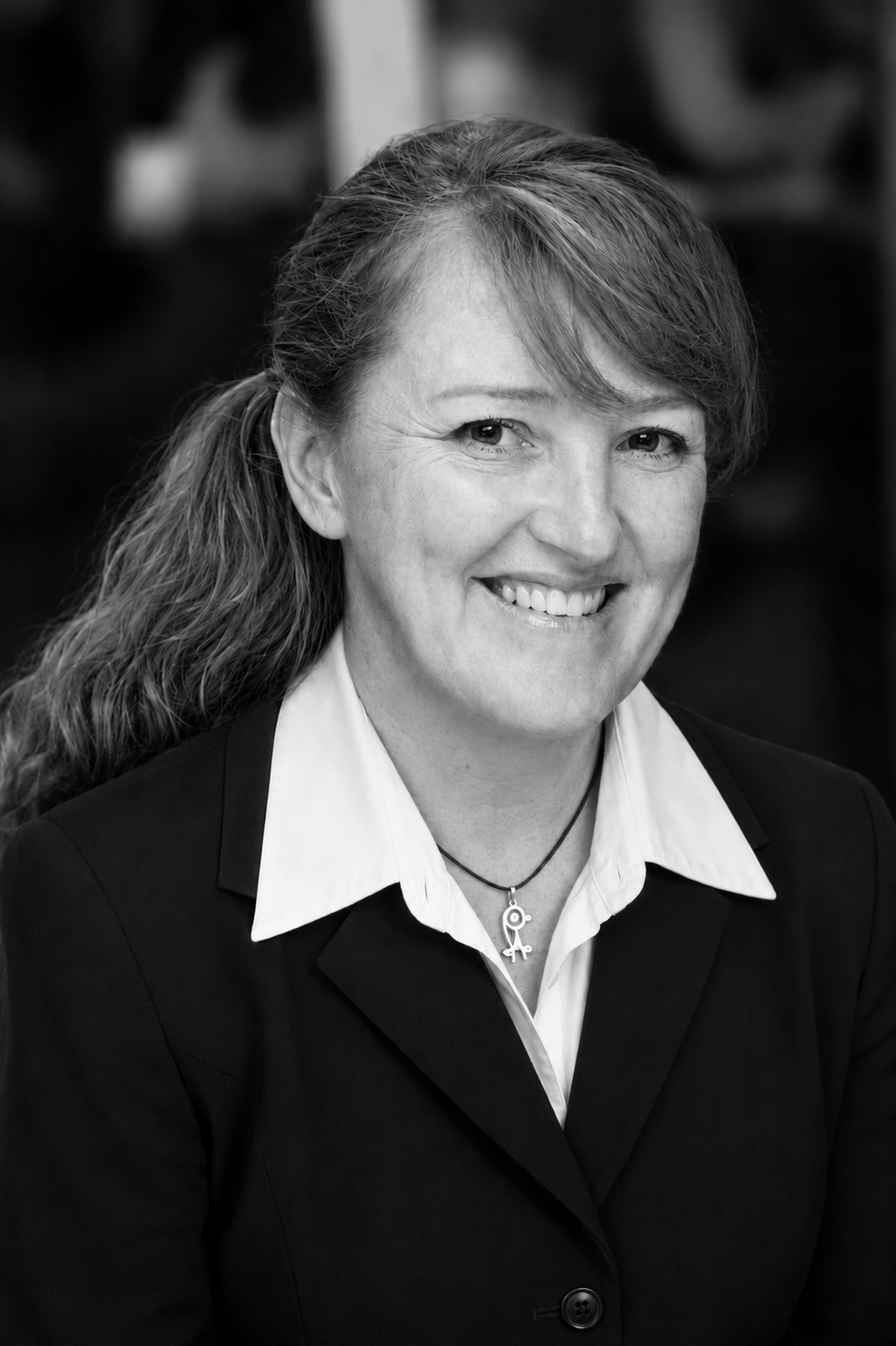 Black and white photo of a smiling woman with long hair tied in a side ponytail, wearing a suit jacket and a necklace with a key pendant.