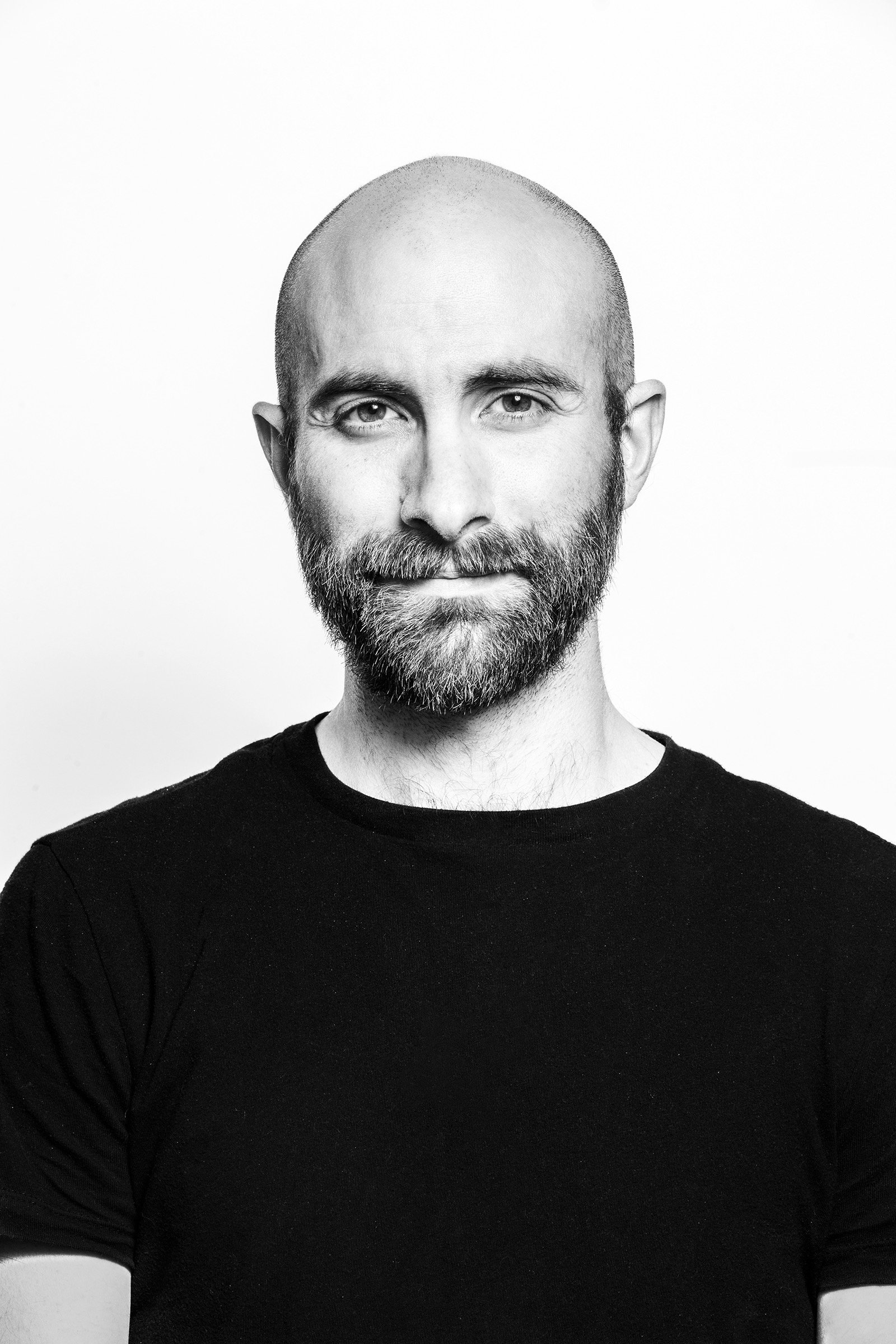 A black and white portrait of a man with a bald head and a full beard, wearing a black T-shirt, against a plain light background.