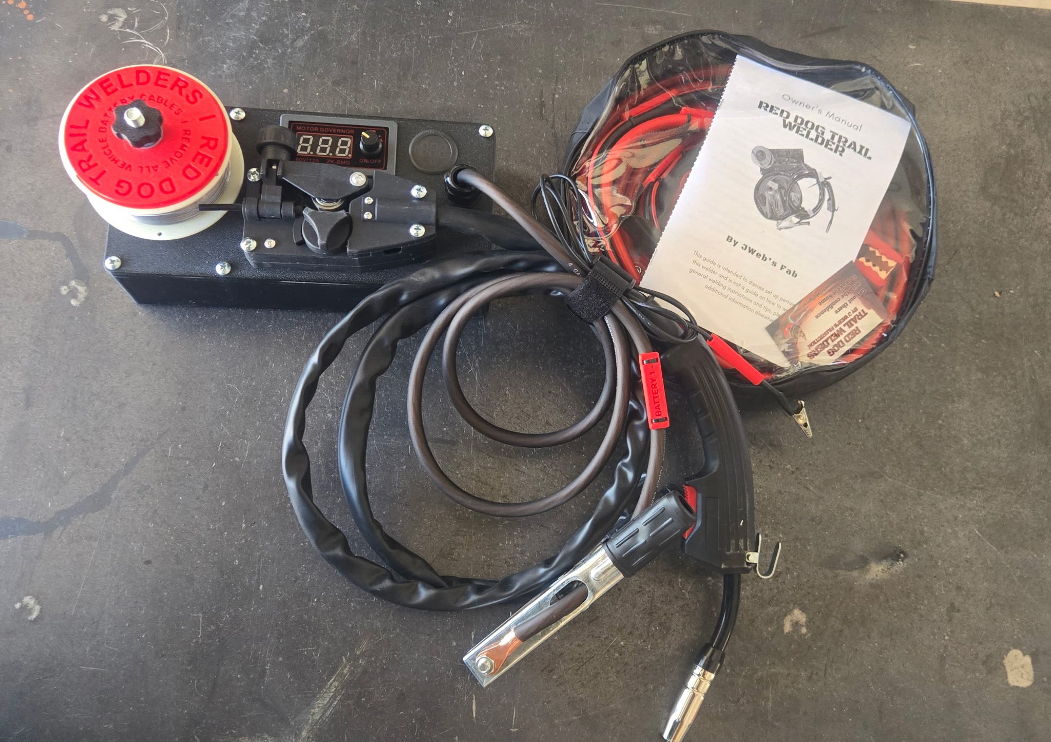 A welding machine with a digital control panel, red welding spool, and various cables and tools on a black surface, alongside a bag of welding accessories labeled 'Reed Dog Trail Welders'.