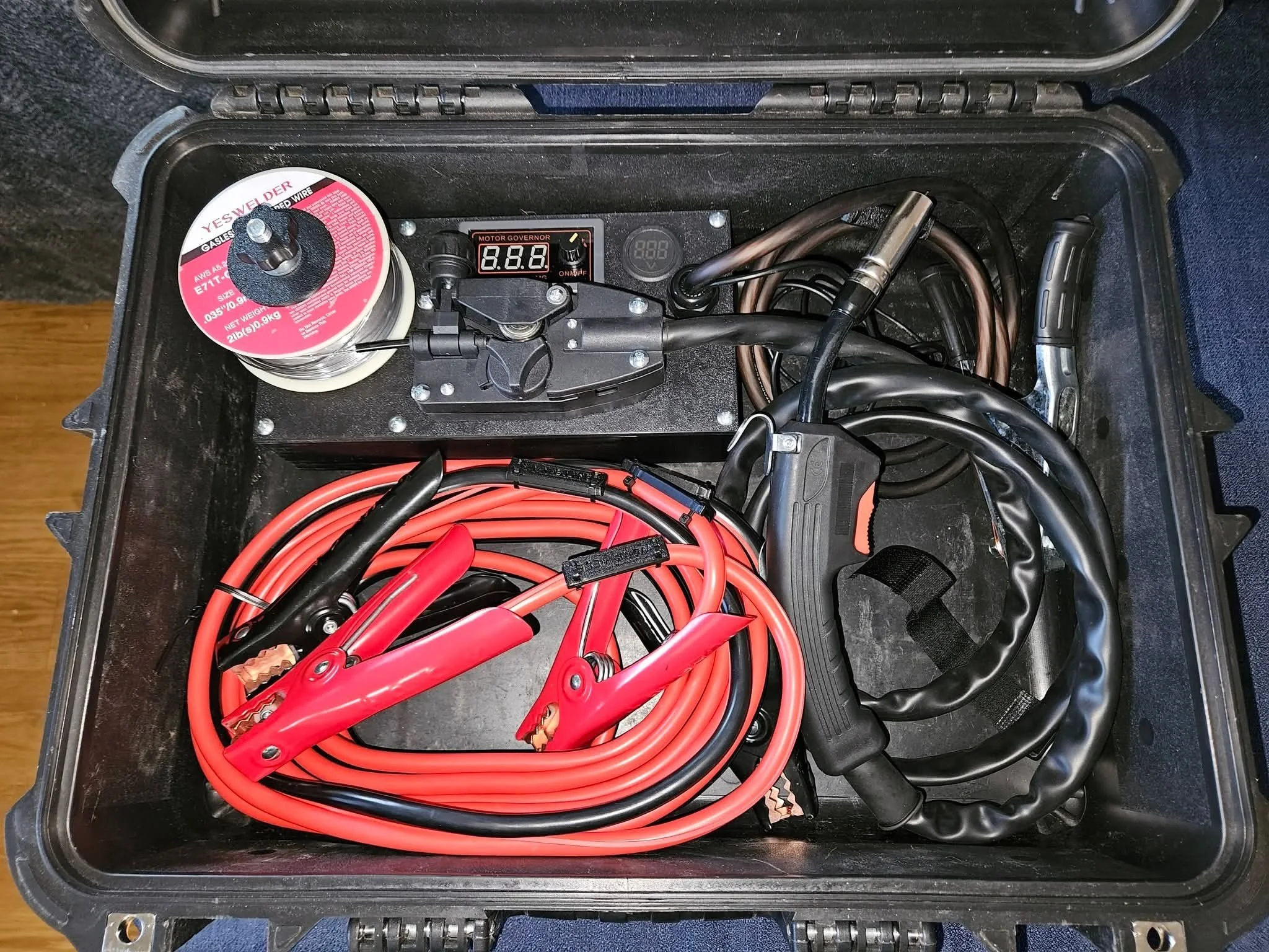 A black plastic case with various electronic components inside, including a spool of wire labeled 'YES WE DER' and a digital motor governor display showing '8.8.8'. Several thick red and black cables are tangled inside the case.
