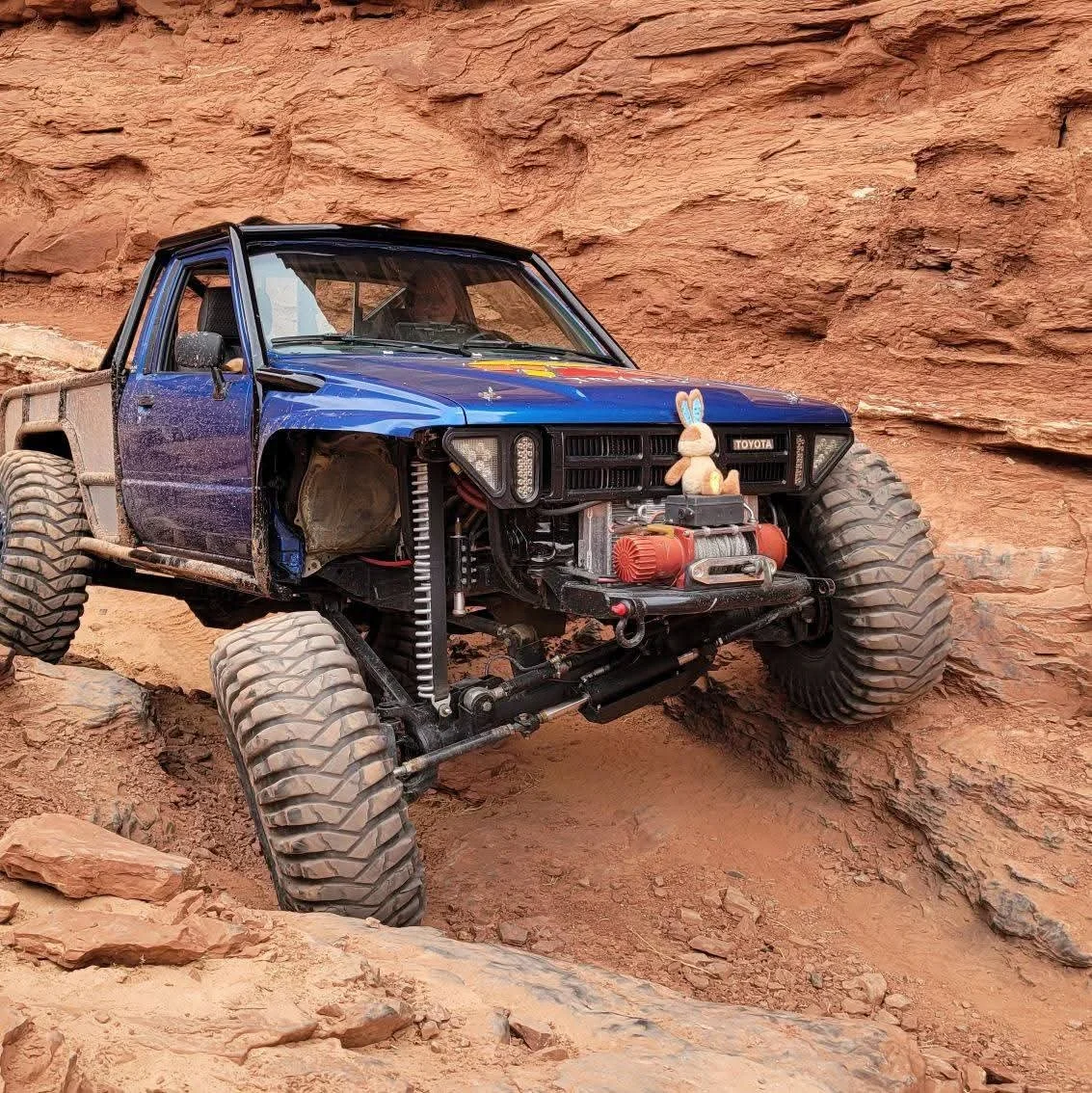 An off-road vehicle with a blue body, large tires, and front missing, navigating on rocky terrain against a red rock wall. A small plush bunny toy sits on the front of the vehicle.