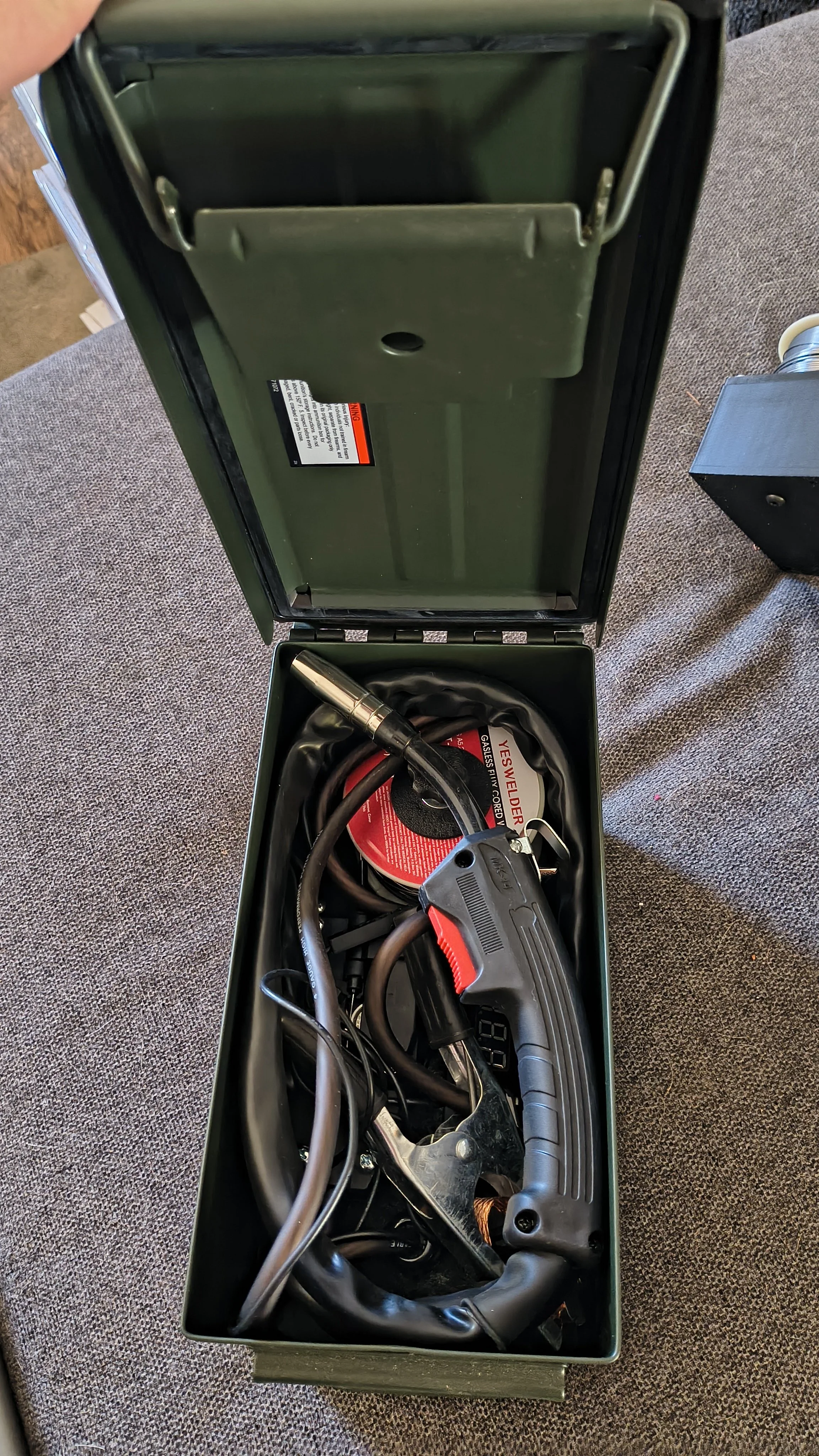 An open metal storage box contains a heated soldering iron with a cord, a spool of solder, and other tools.