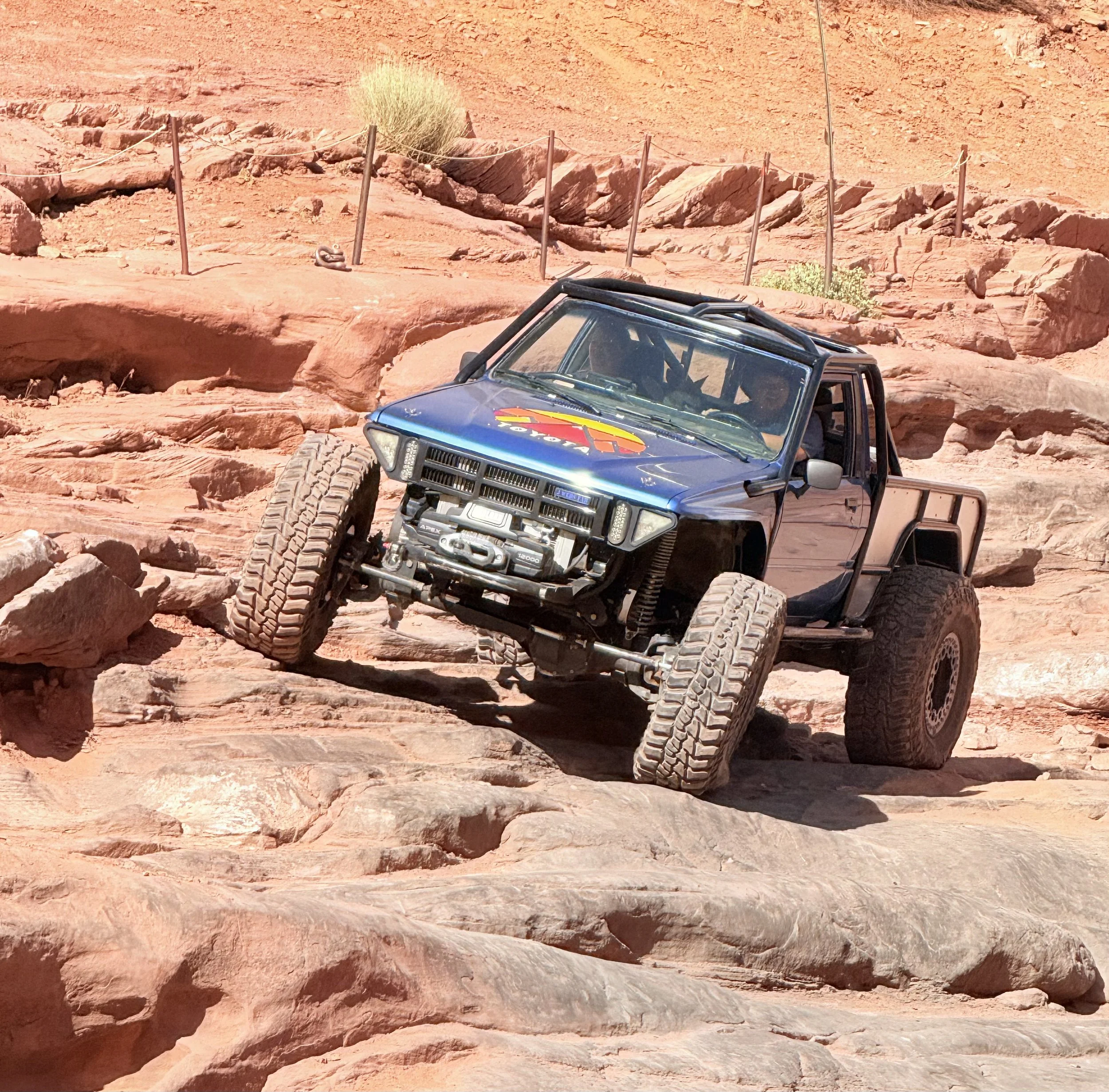 A blue off-road vehicle with a colorful logo on the hood driving over a rocky, desert terrain with orange sand and rock formations.