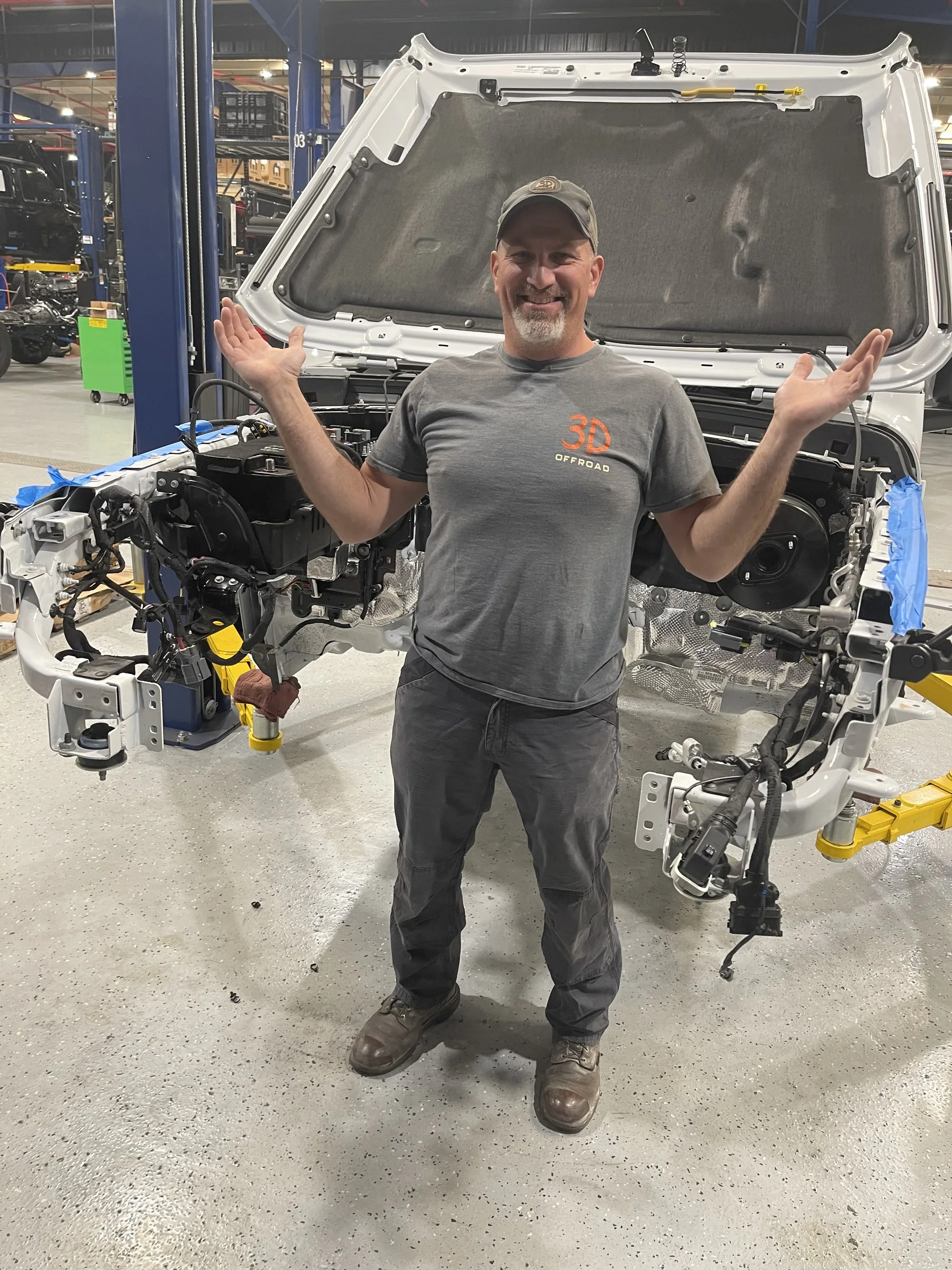 A man wearing a cap and gray t-shirt standing inside an industrial workshop with a partially assembled vehicle frame and a car lift in the background, smiling and posing with arms raised.