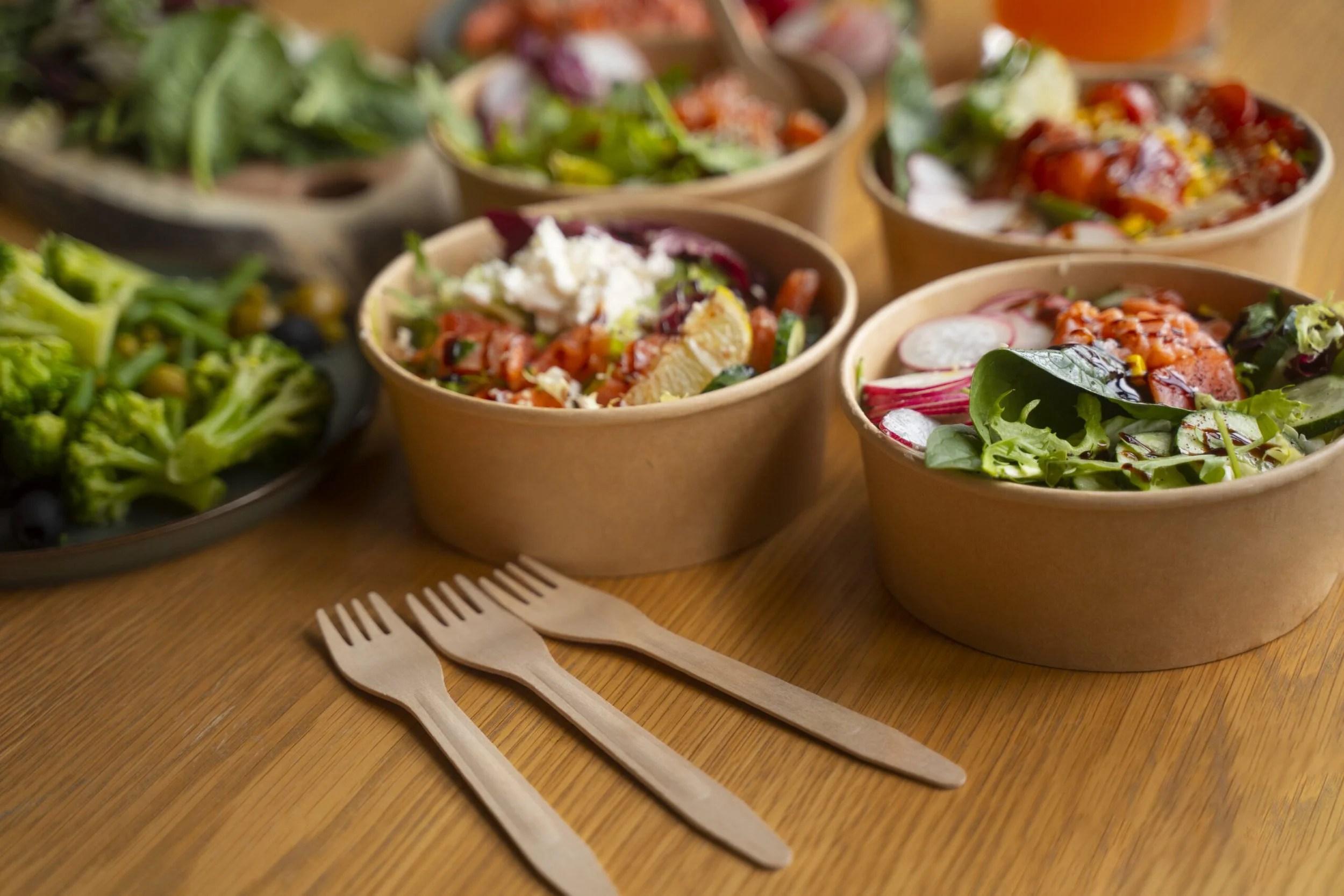 Various bowls of colorful salads and greens on a wooden table with two wooden forks in the foreground.