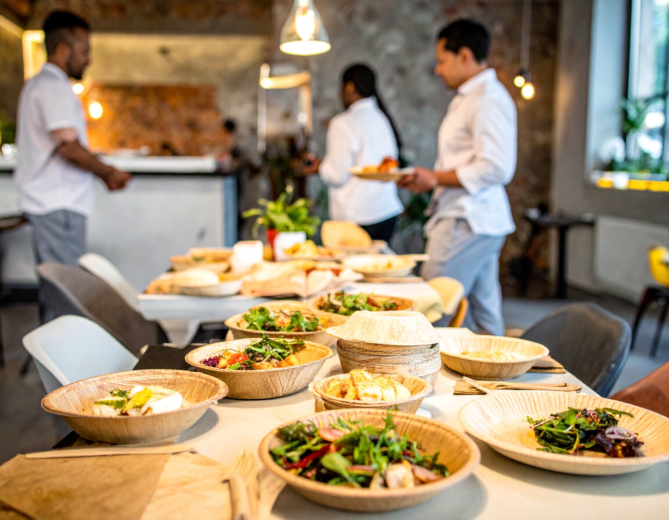A table displays a variety of dishes served in edible bowls, with several people in the background at a buffet-style event, in a rustic setting with exposed brick walls and hanging lights.