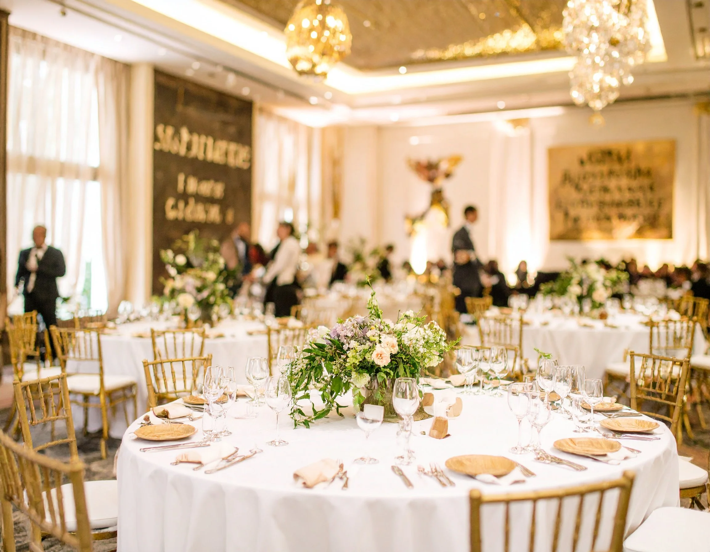 Elegant banquet hall set up for a wedding reception with round tables, gold chairs, floral centerpieces, and chandeliers.
