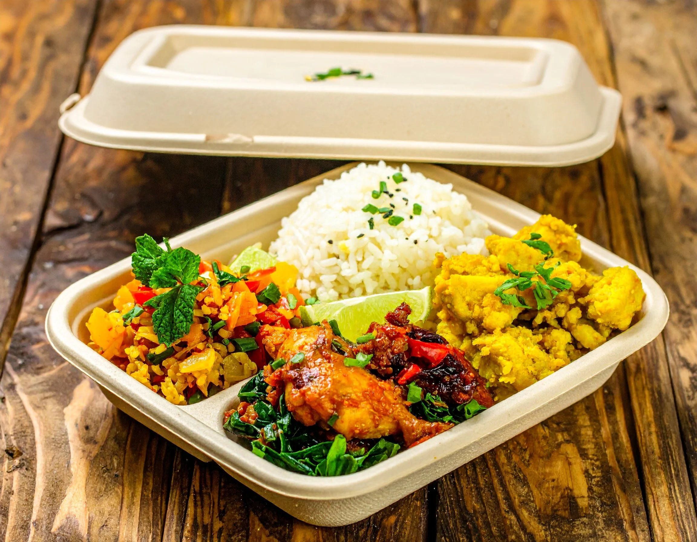 Takeout container on a wooden surface filled with rice, vegetables, chicken, and garnished with herbs and lime.