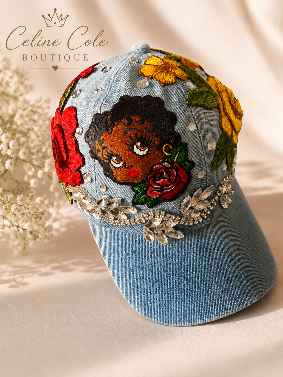 Blue denim baseball cap decorated with embroidered flowers, rhinestones, and a patch of a woman's face with curly hair and red lipstick, displayed against a neutral background with white flowers.