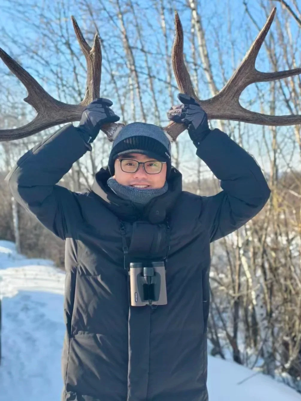 Man outdoors in winter clothing holding large elk antlers above his head, smiling, with snow-covered ground and bare trees in the background.