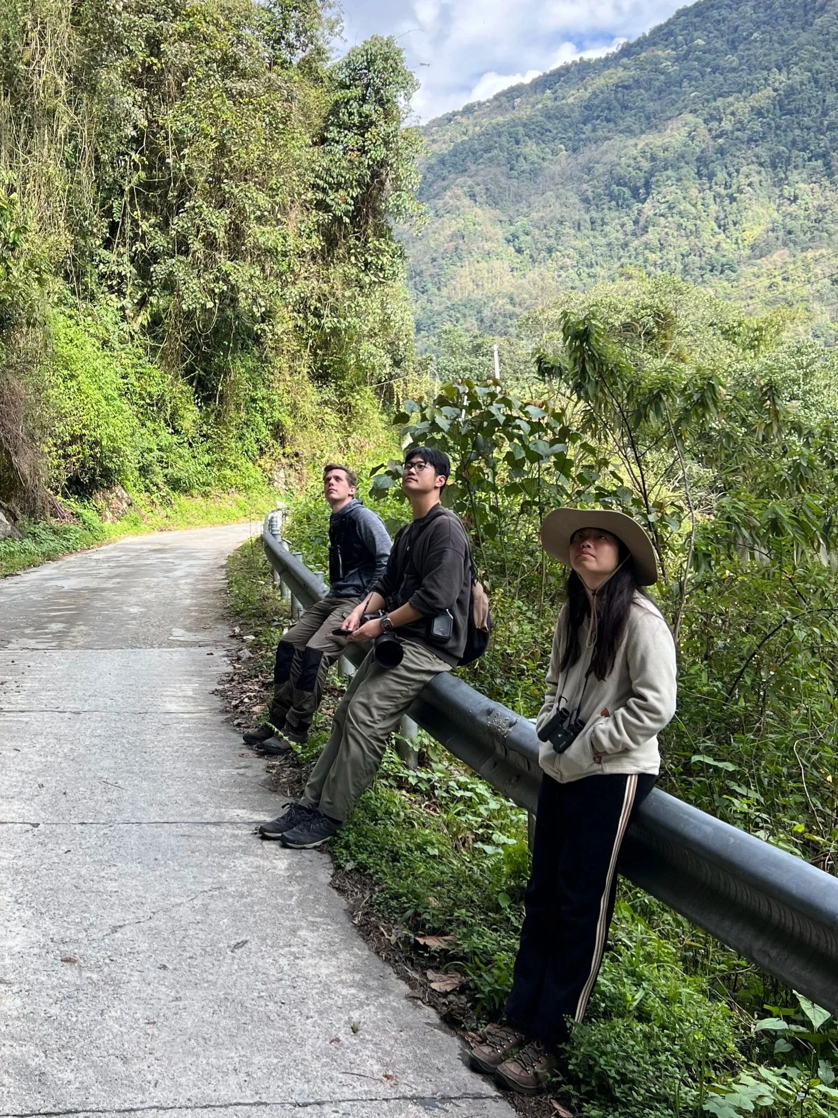 Three hikers resting along a mountain trail with lush green vegetation and a scenic mountain view in the background.