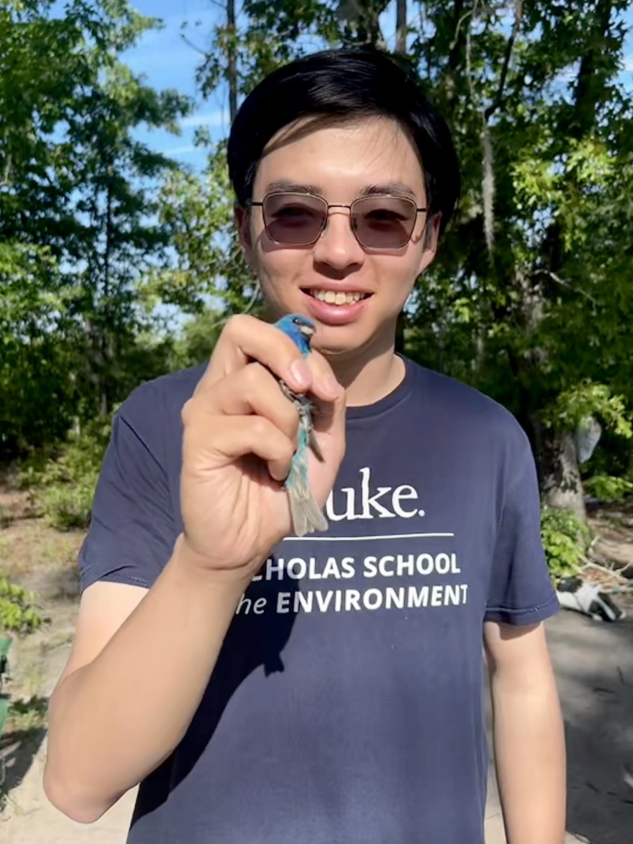 Young person wearing sunglasses, holding a small blue bird, outdoors with trees and blue sky in the background. They are smiling and wearing a navy T-shirt with partially visible white text.