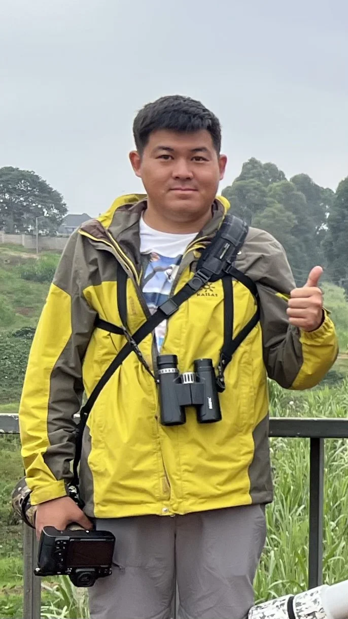 Man standing outdoors in a yellow and gray jacket, carrying binoculars and a camera, and giving a thumbs-up.