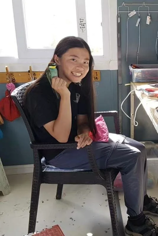 A girl sitting on a black chair in a classroom, holding a green and yellow toy, smiling at the camera. There is a window, a whiteboard with hooks, and school supplies in the background.