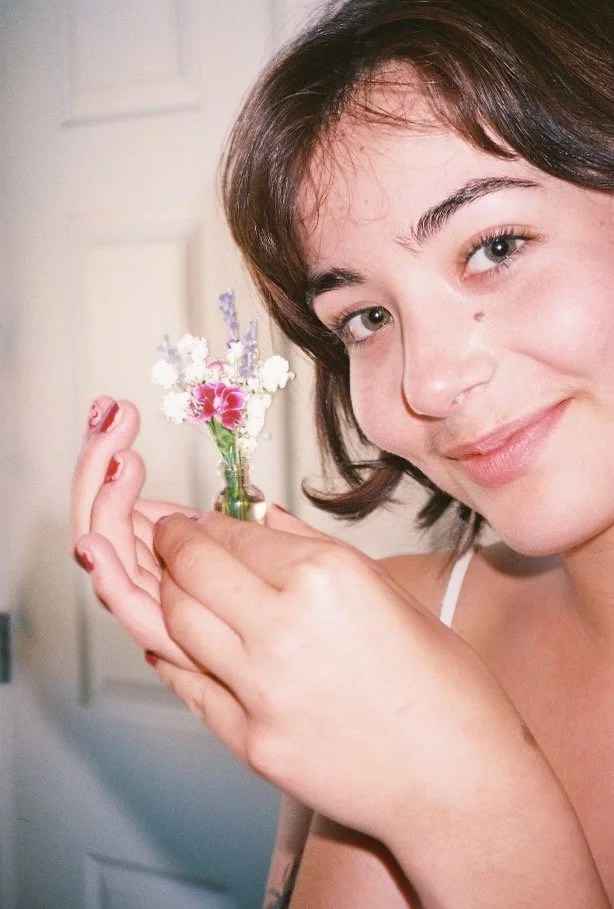 A young woman with short brown hair holding a small glass vase with pink and white flowers, smiling at the camera.