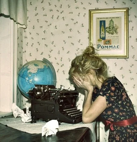 A woman with blonde hair tied in a bun sitting at a table in a vintage-style room, covering her face with her hands while facing a vintage typewriter.