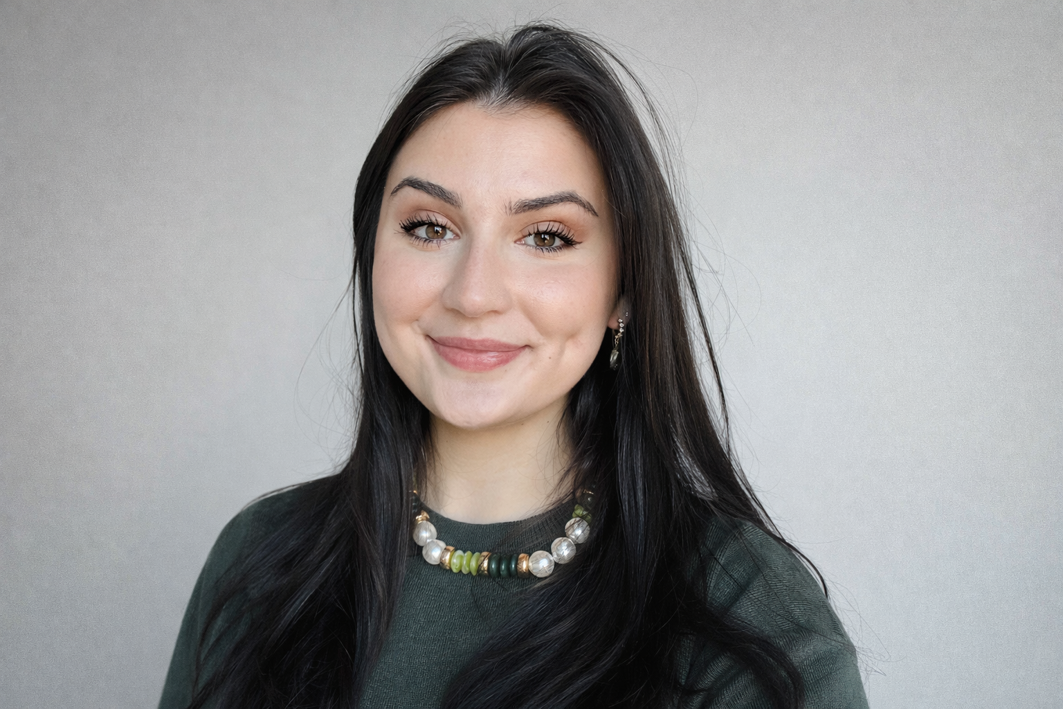A young woman with long dark hair, smiling, wearing a green top, a pearl and green beaded necklace, and earrings, standing against a plain light background.