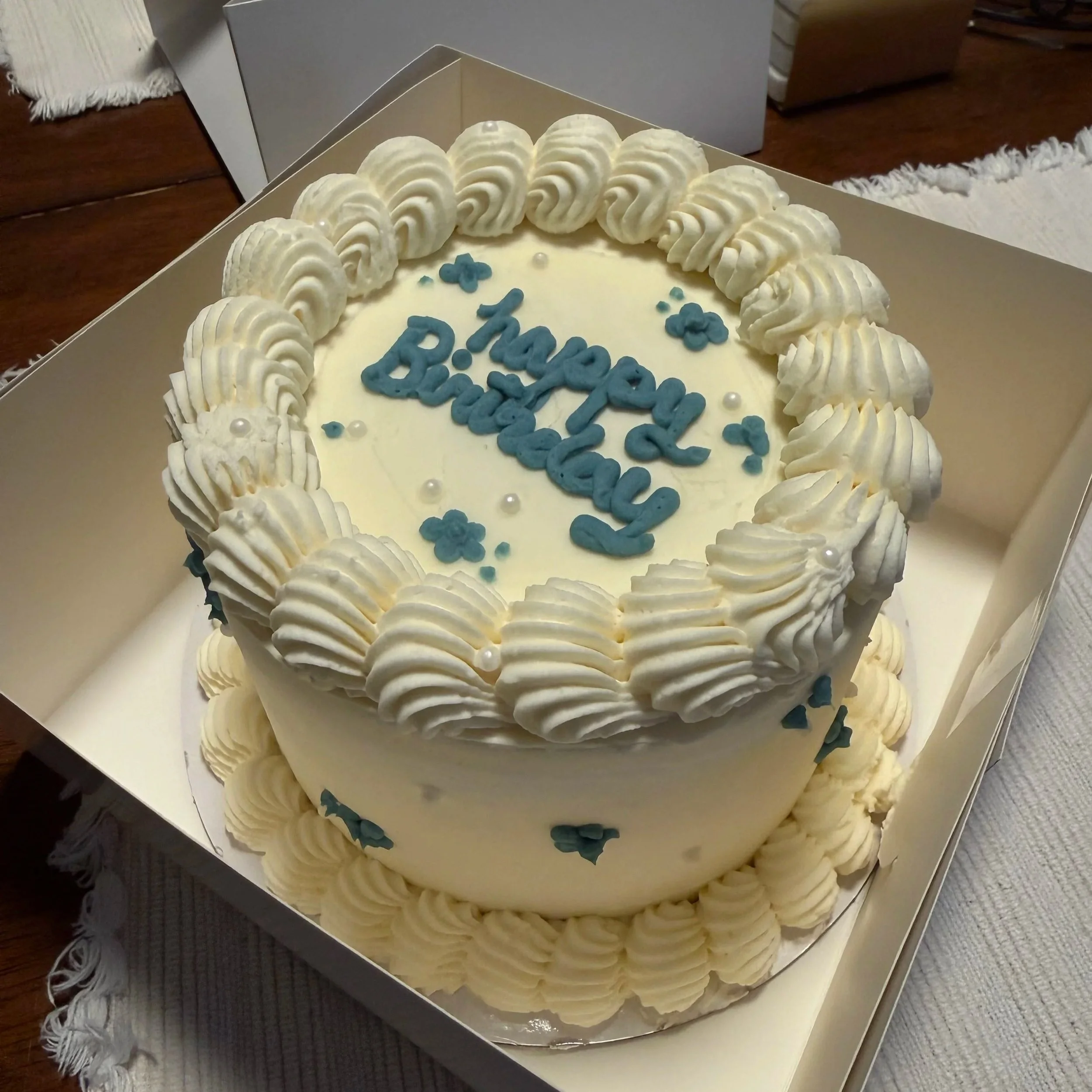 A decorated birthday cake with white frosting and piped cream border, with blue icing letters and small decorative elements, placed in an open box on a table.