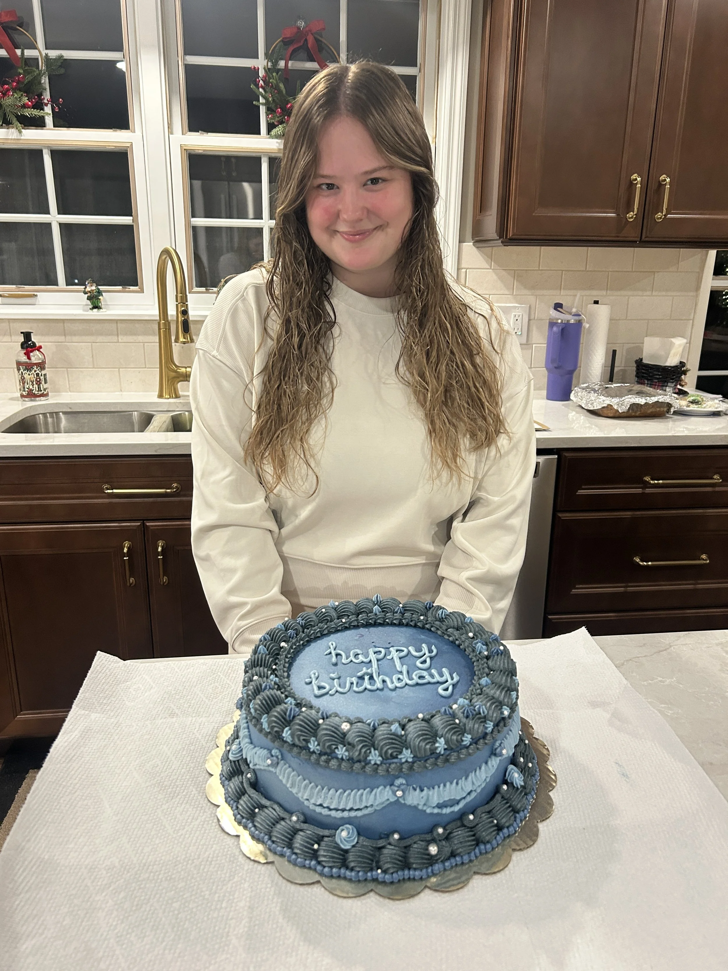 A woman with long, wavy hair and a white sweatshirt standing behind a birthday cake with blue frosting, decorated with piped swirls and silver beads, with a cake topper that says 'happy birthday', in a kitchen with wooden cabinets and holiday decorat