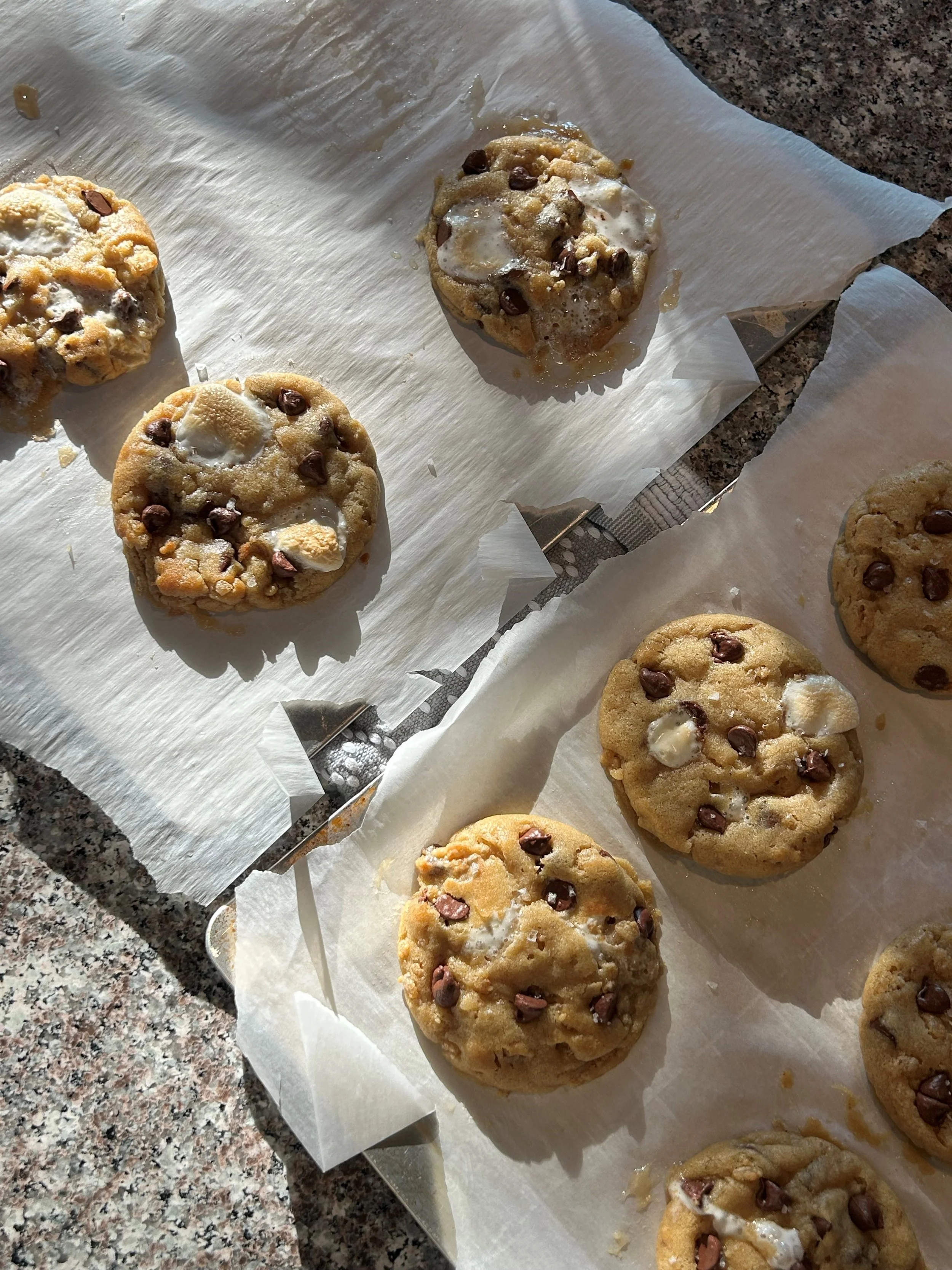 Several chocolate chip cookies on parchment paper, some with melted chocolate and white chocolate chips, on a speckled granite countertop.