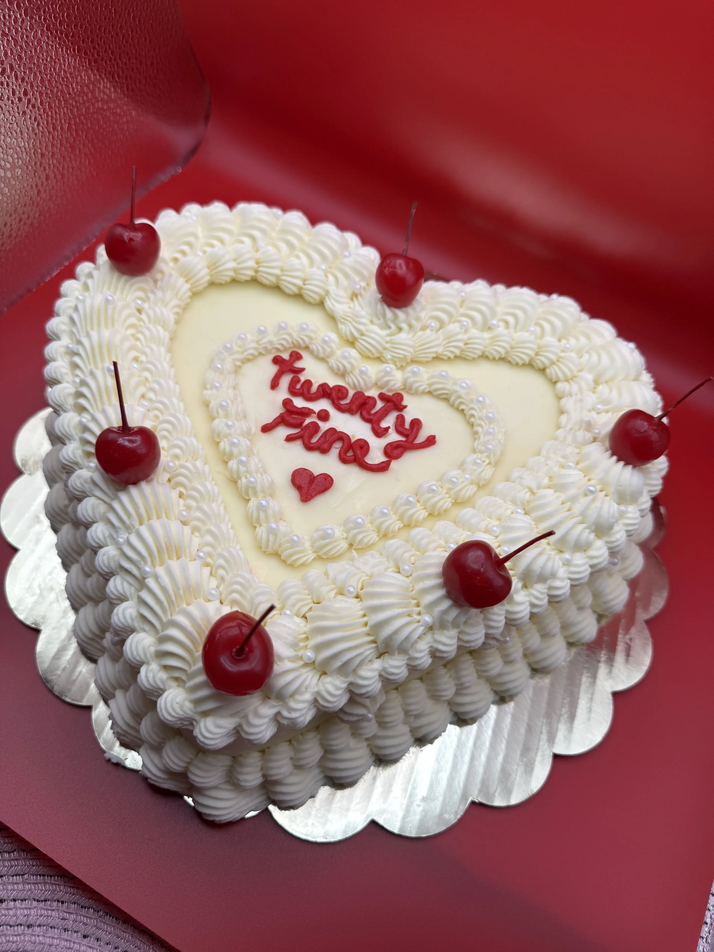 Round birthday cake decorated with white frosting, red cherries on top, and a message that reads 'Happy birthday Timi' in red icing, with a red heart underneath.