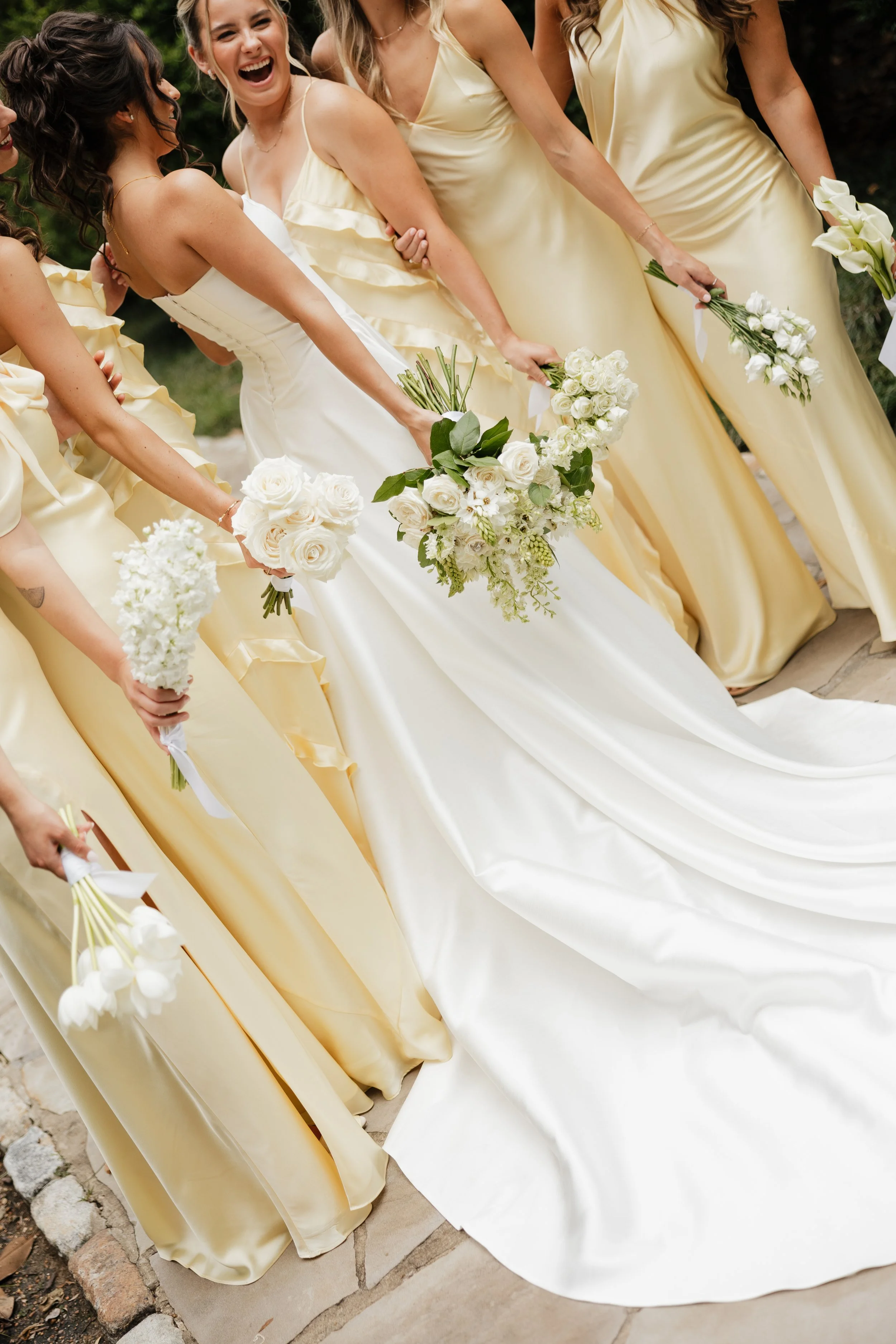 Group of women in yellow dresses holding bouquets, standing outdoors, smiling and laughing, wedding celebration.