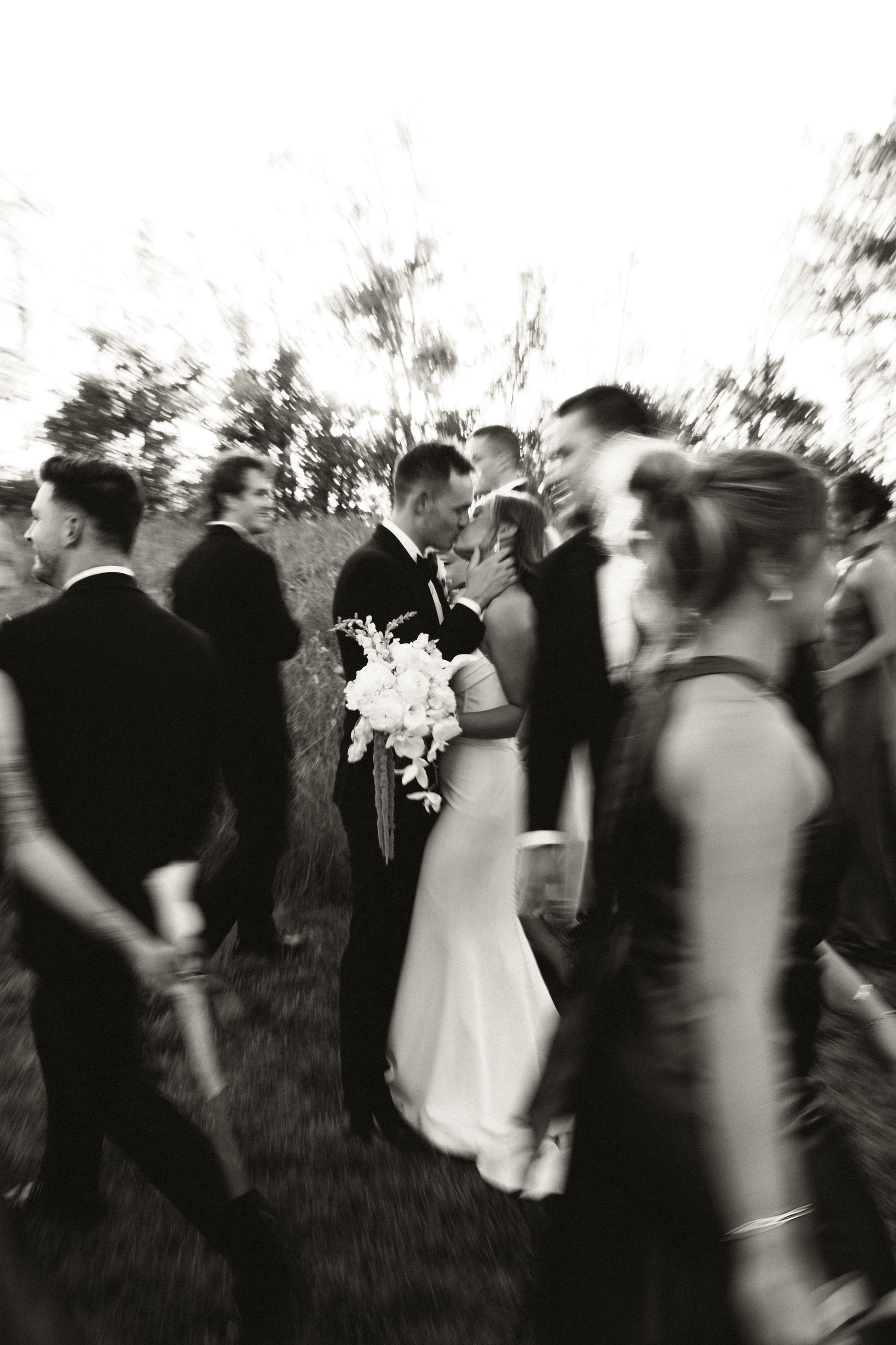 A black and white photo of people at a wedding, with a couple kissing surrounded by guests, trees, and an outdoor setting.