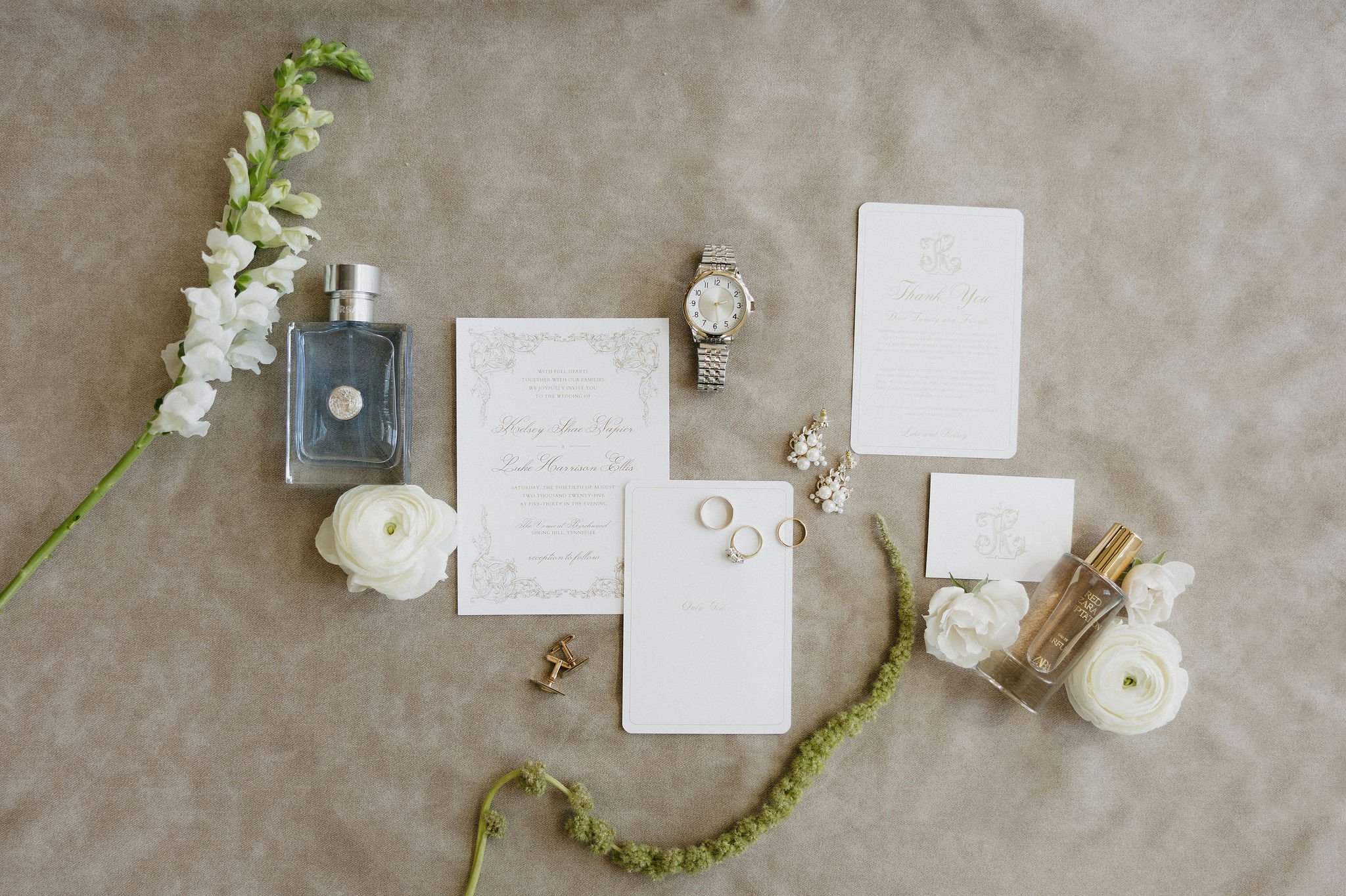 Wedding invitation suite, jewelry, flowers, and perfume bottles arranged on a beige surface.