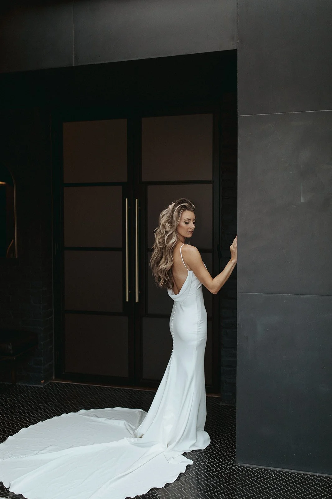 A woman in a white wedding gown standing indoors against a black door and dark walls, with her back partially turned and gazing downward.