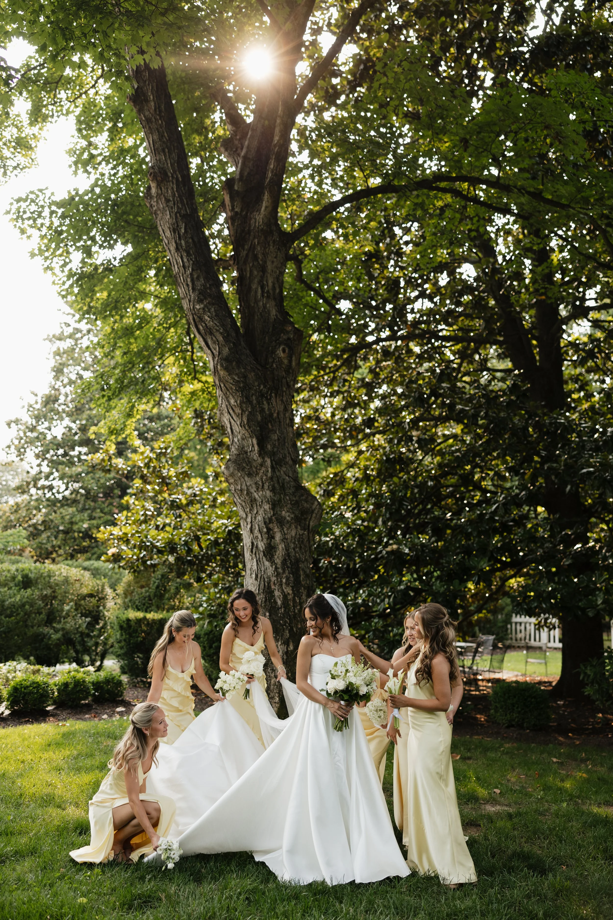 A group of women in yellow dresses helping a bride in a white wedding gown outdoors under a large tree with green leaves and sunlight shining through. The bride is holding a bouquet of white flowers.