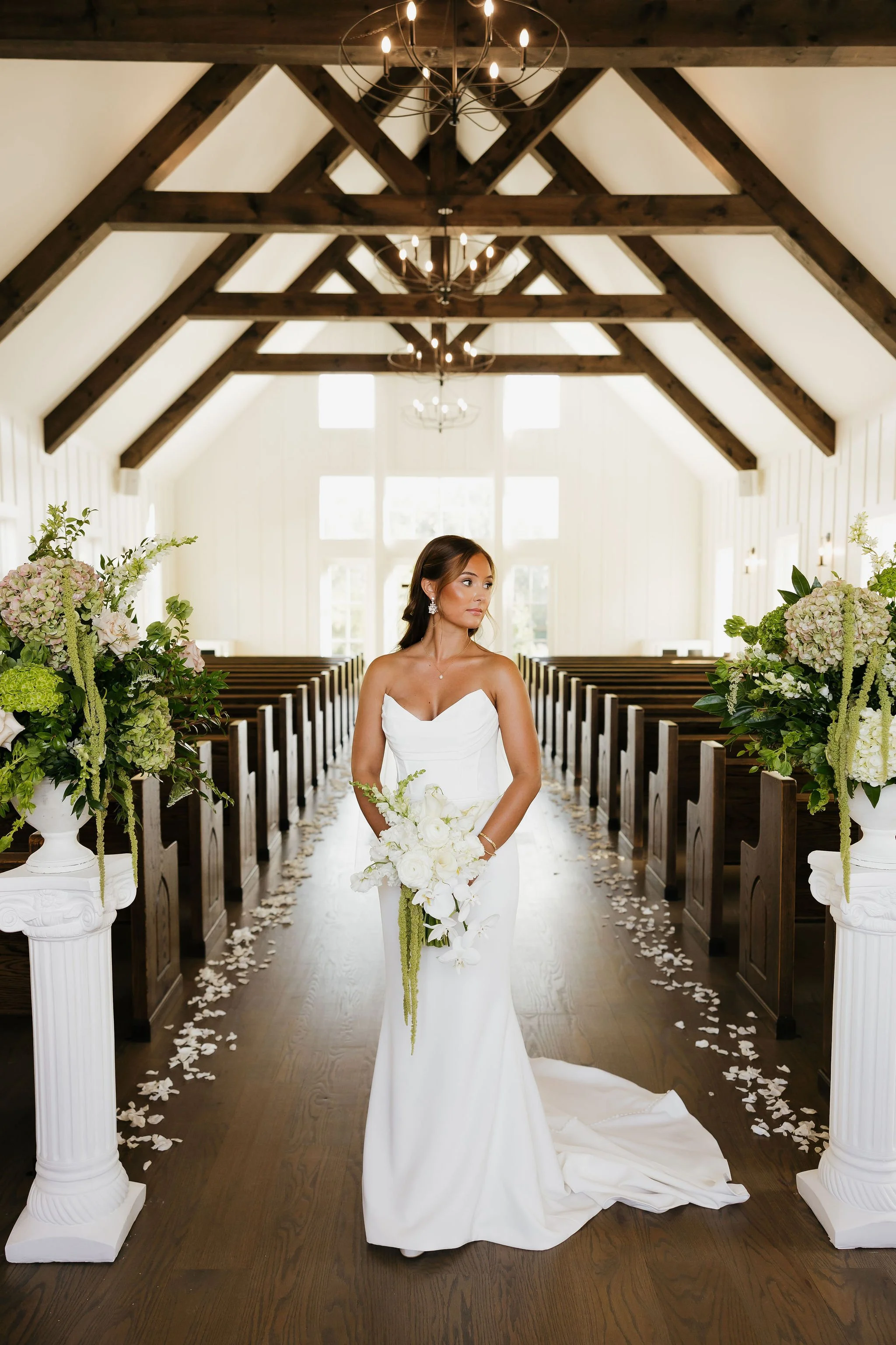 A bride in a white wedding dress standing in a church aisle, holding a bouquet, with flower arrangements on either side and wooden beams on the ceiling.