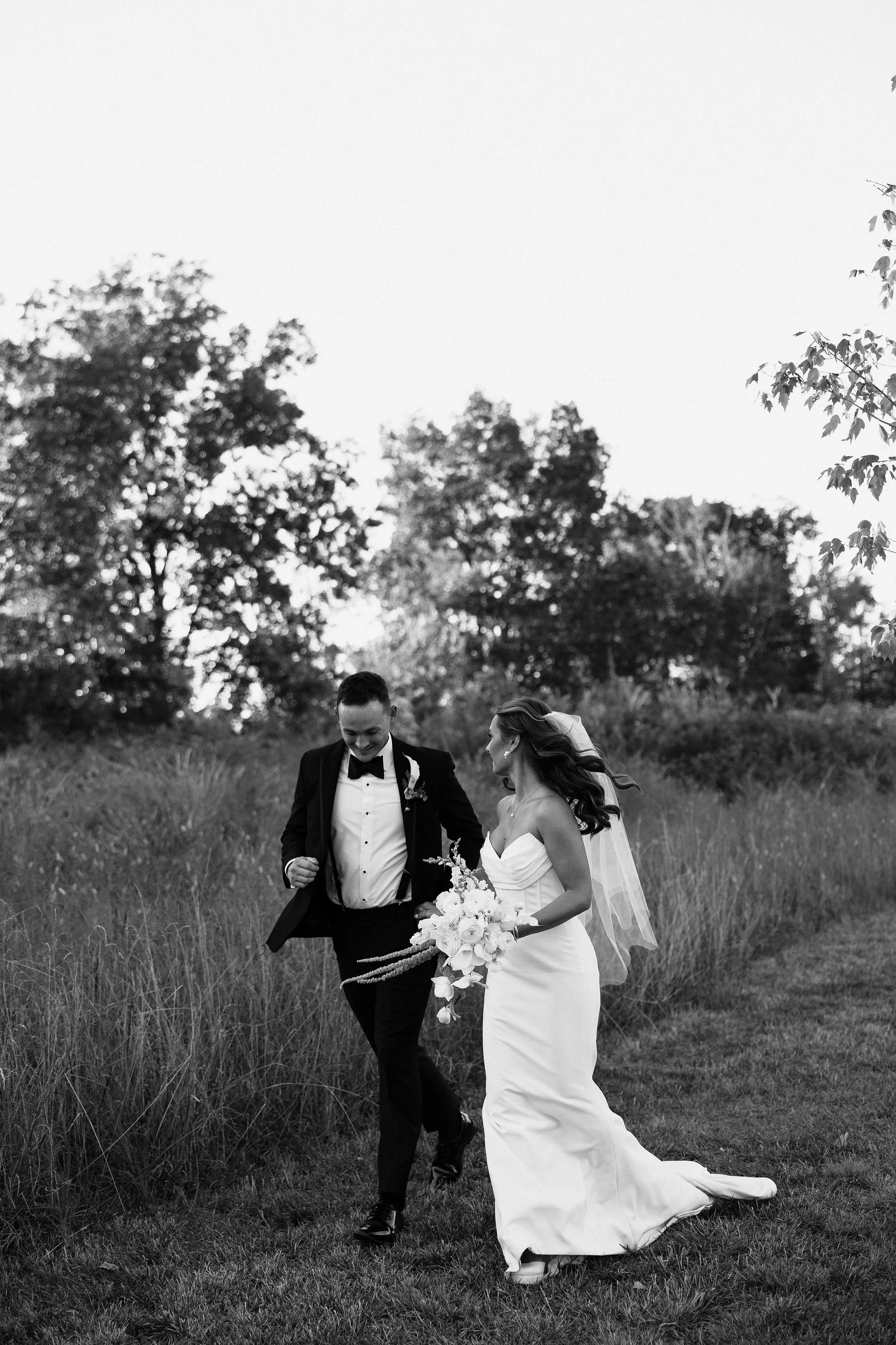 A black and white photo of a newlywed couple walking through a grassy field, holding hands and smiling, with trees in the background.
