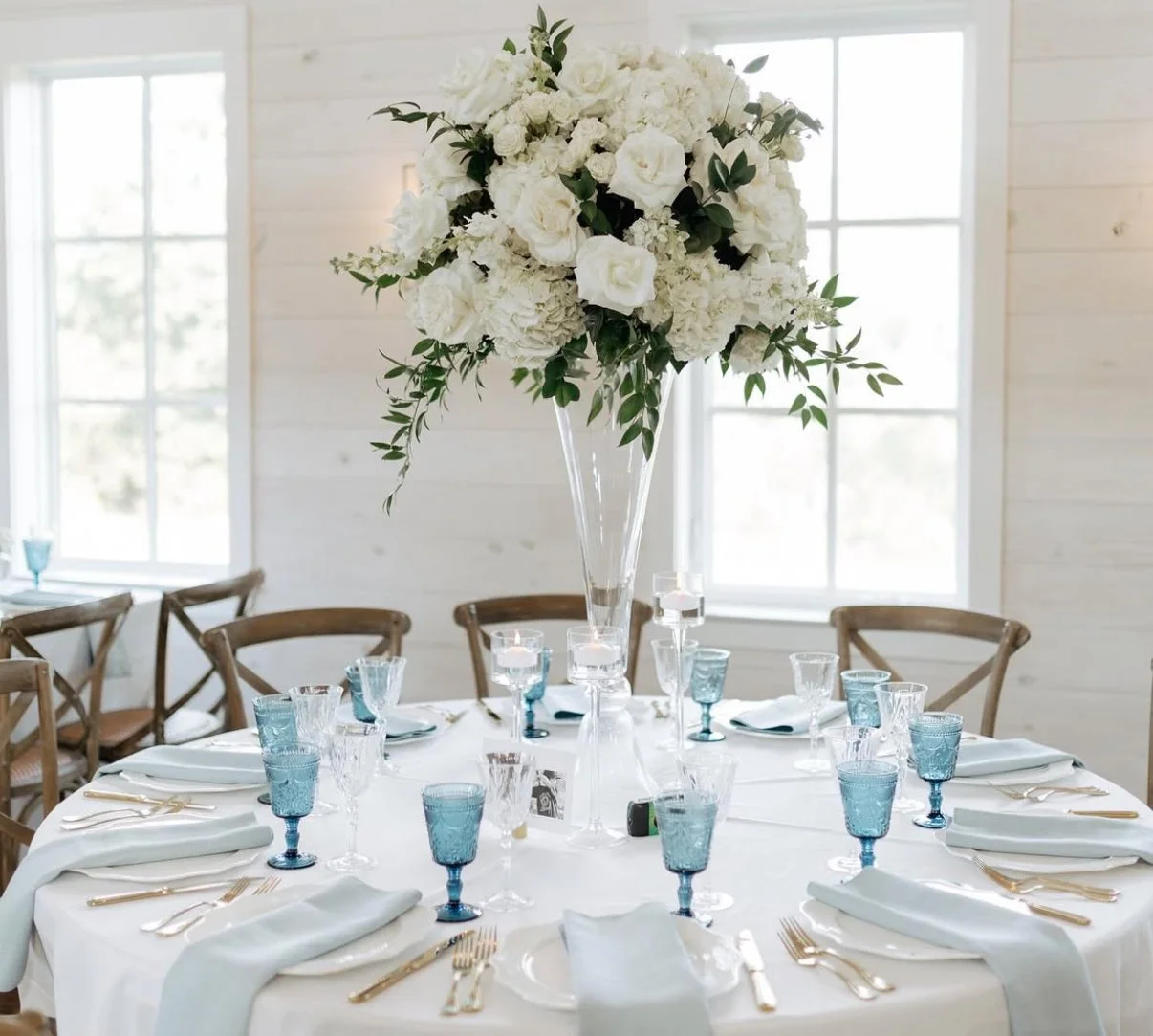 A round wedding table with white tablecloth, set with gold utensils, white napkins, and turquoise glassware, topped with a large floral centerpiece of white roses and greenery in a tall glass vase, in a bright room with wooden chairs and multiple large windows.