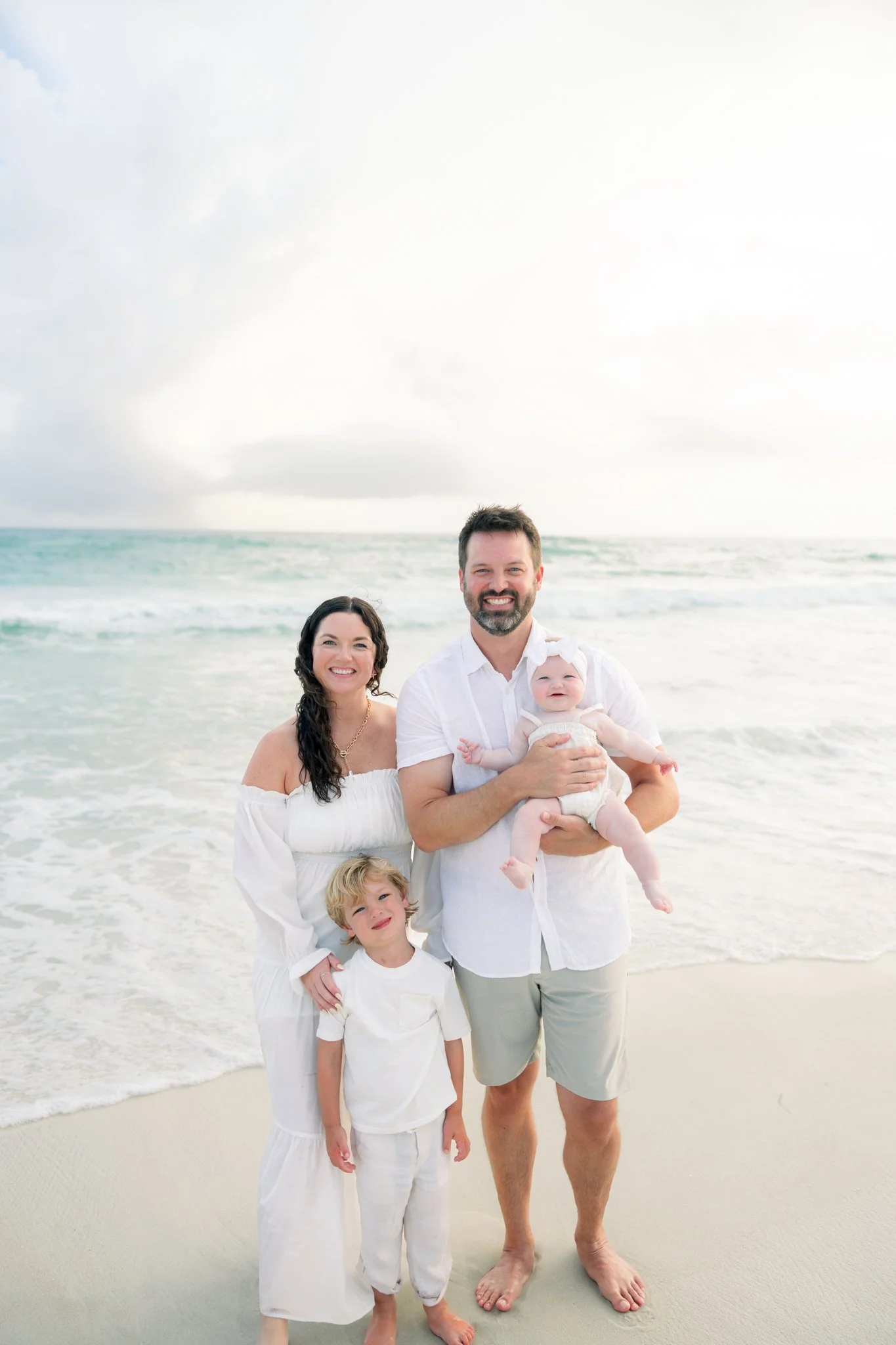 Family of five at the beach, standing in shallow water with waves and cloudy sky in the background, smiling and dressed in white summer clothing.