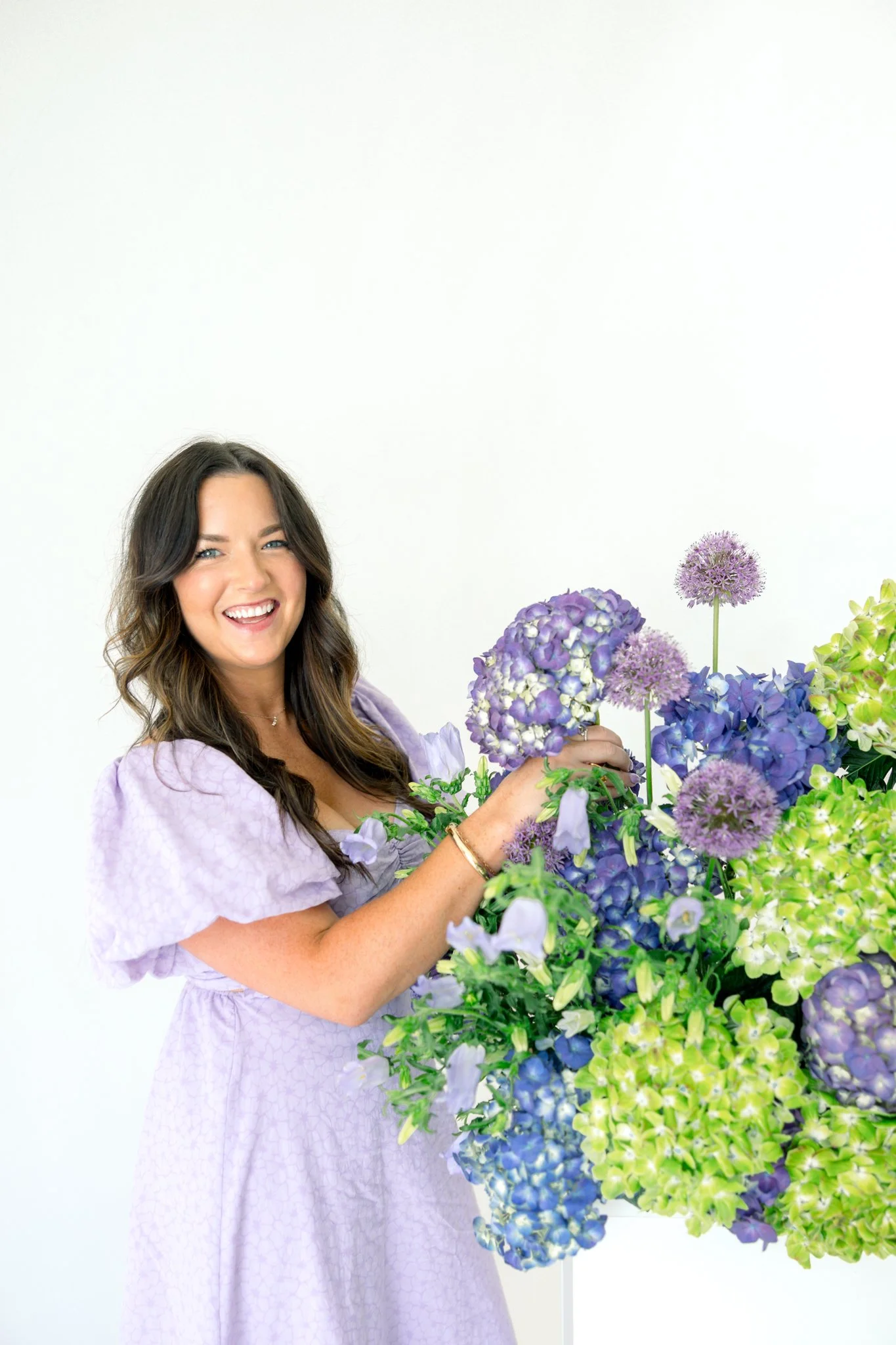 A woman with long brown hair wearing a light purple dress stands next to a large bouquet of purple and green hydrangea flowers, smiling at the camera.