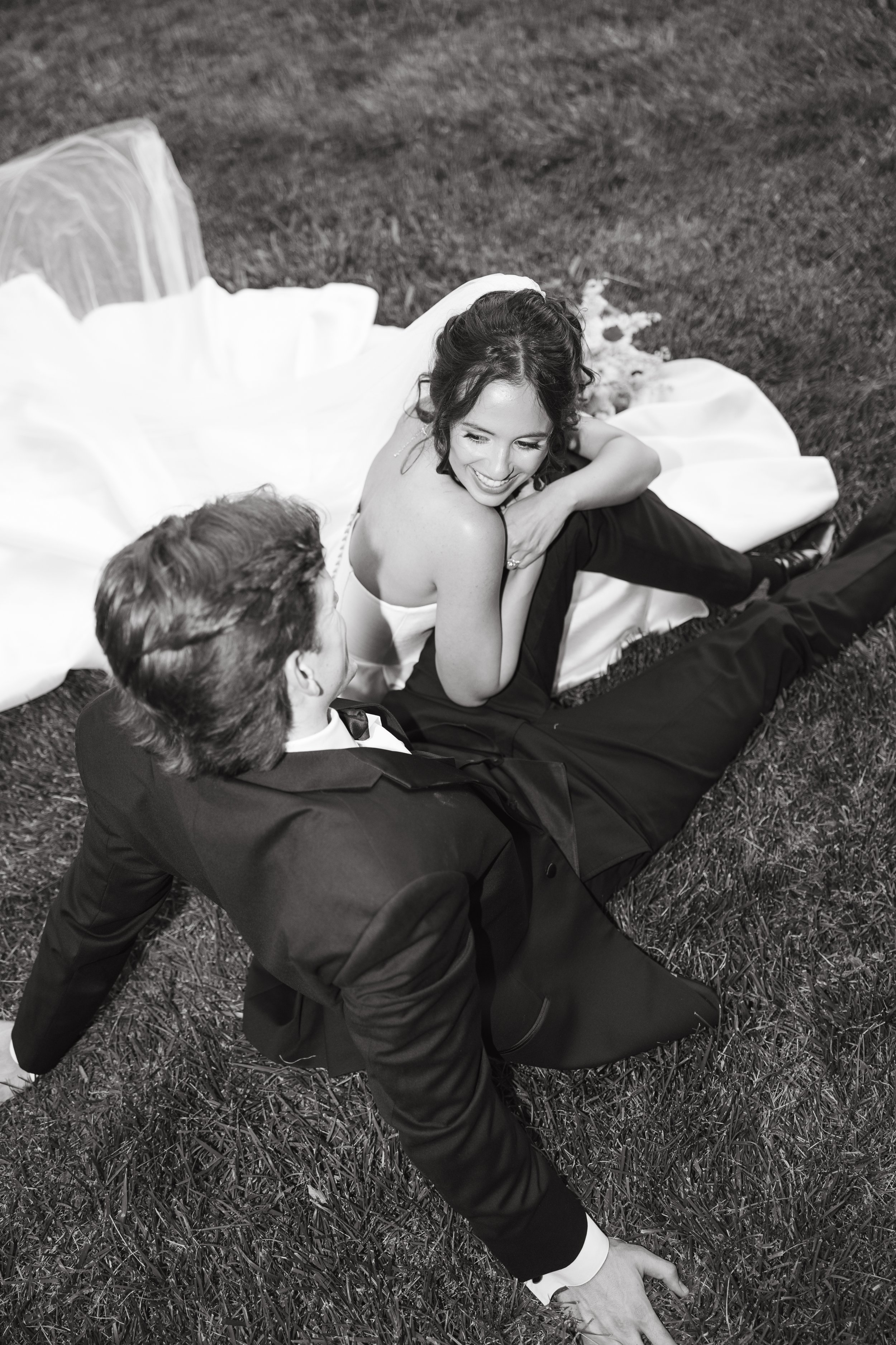 A woman in a wedding dress and a man in a tuxedo sitting on grass, smiling and engaged in a conversation.