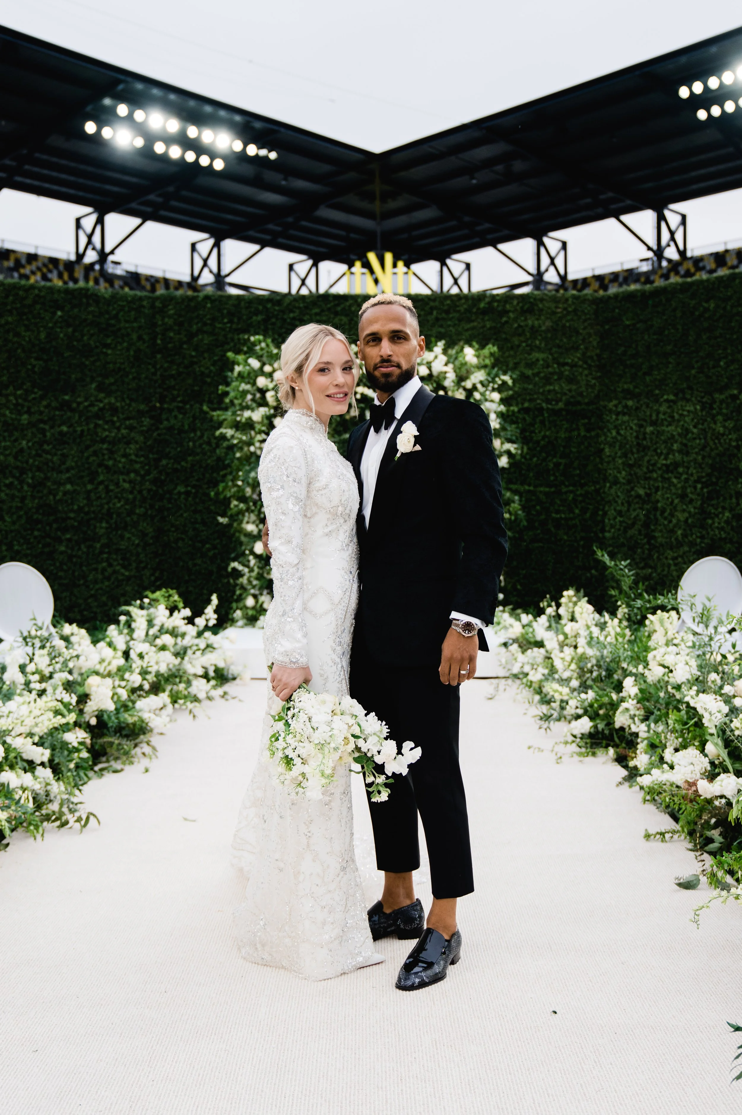 A bride and groom standing together at their wedding, surrounded by white flowers and greenery, with a modern outdoor setting and stadium-like seating in the background.