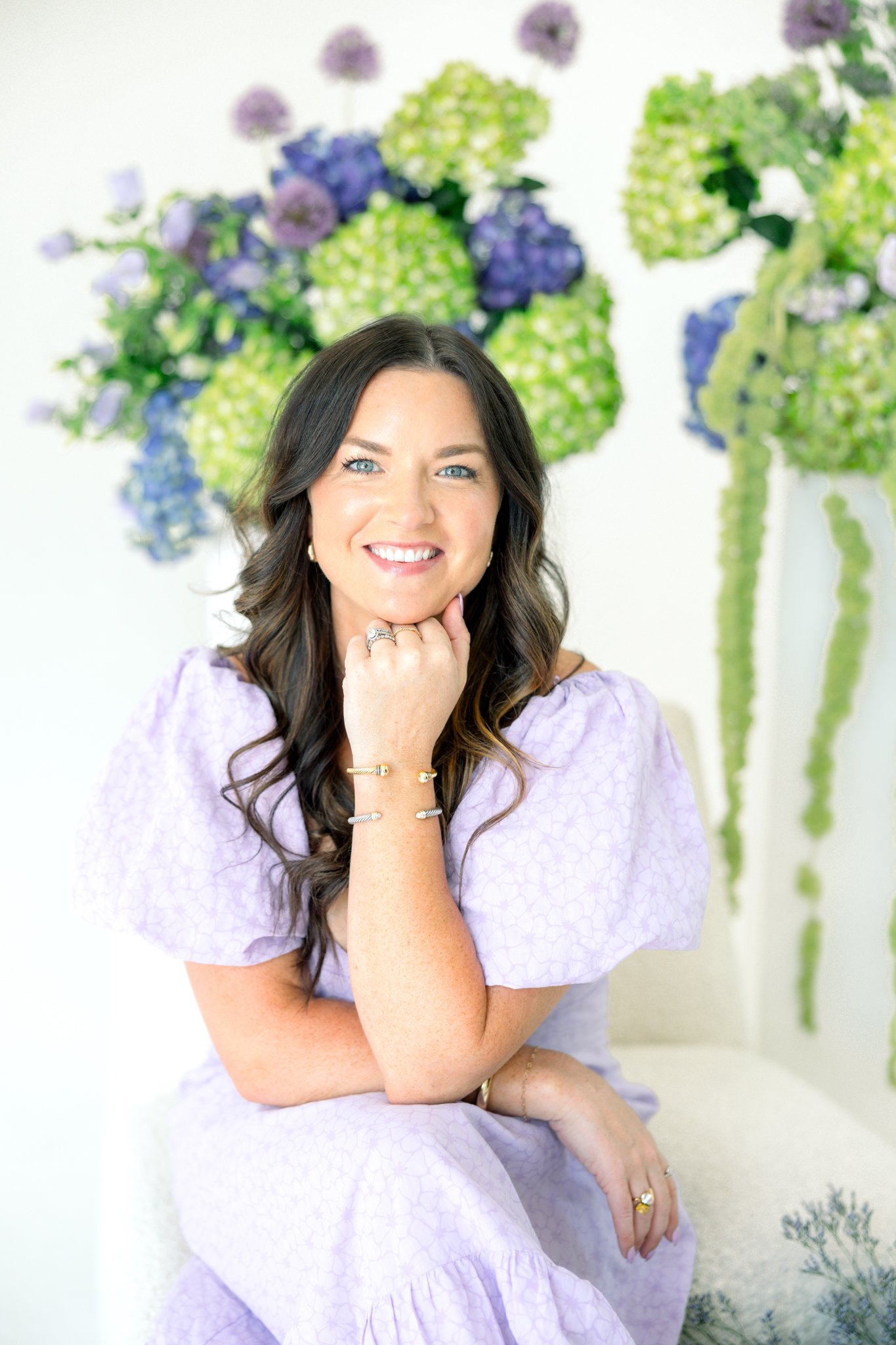 A woman with long brown hair and blue eyes smiling and resting her chin on her hand, wearing a lavender dress with a ruffled sleeve. She is sitting indoors in front of a colorful floral wallpaper featuring green and purple flower arrangements.