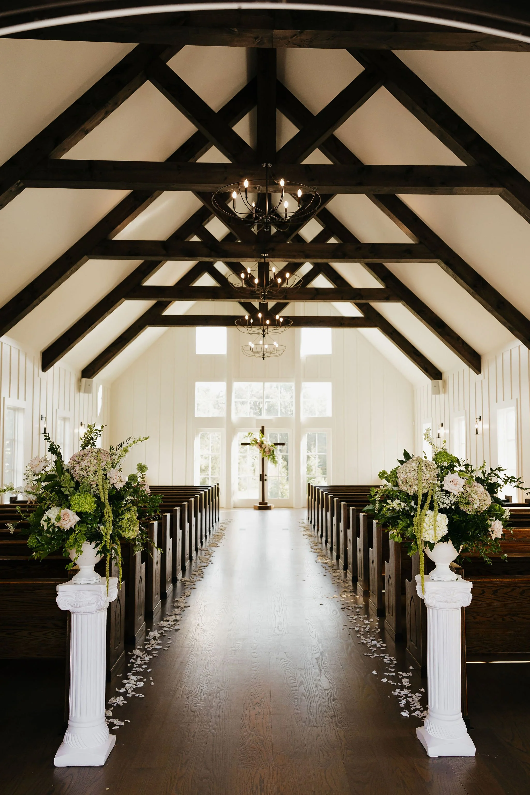 Interior of a church aisle decorated for a wedding, with white flower arrangements on white pedestals and a chandelier hanging from a dark wooden vaulted ceiling.