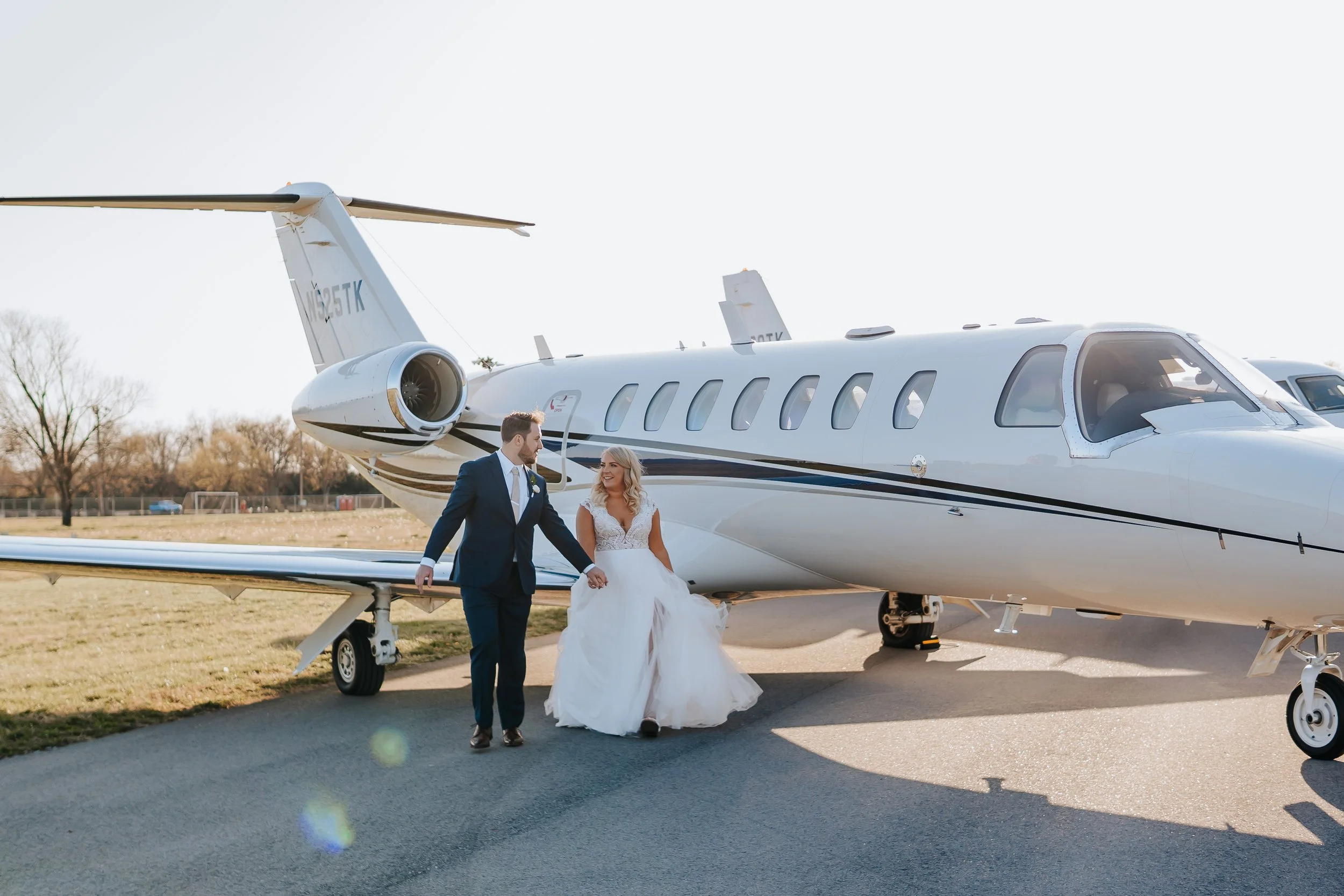 A bride and groom holding hands in front of a small private jet on an airport tarmac, with clear sky and trees in the background.