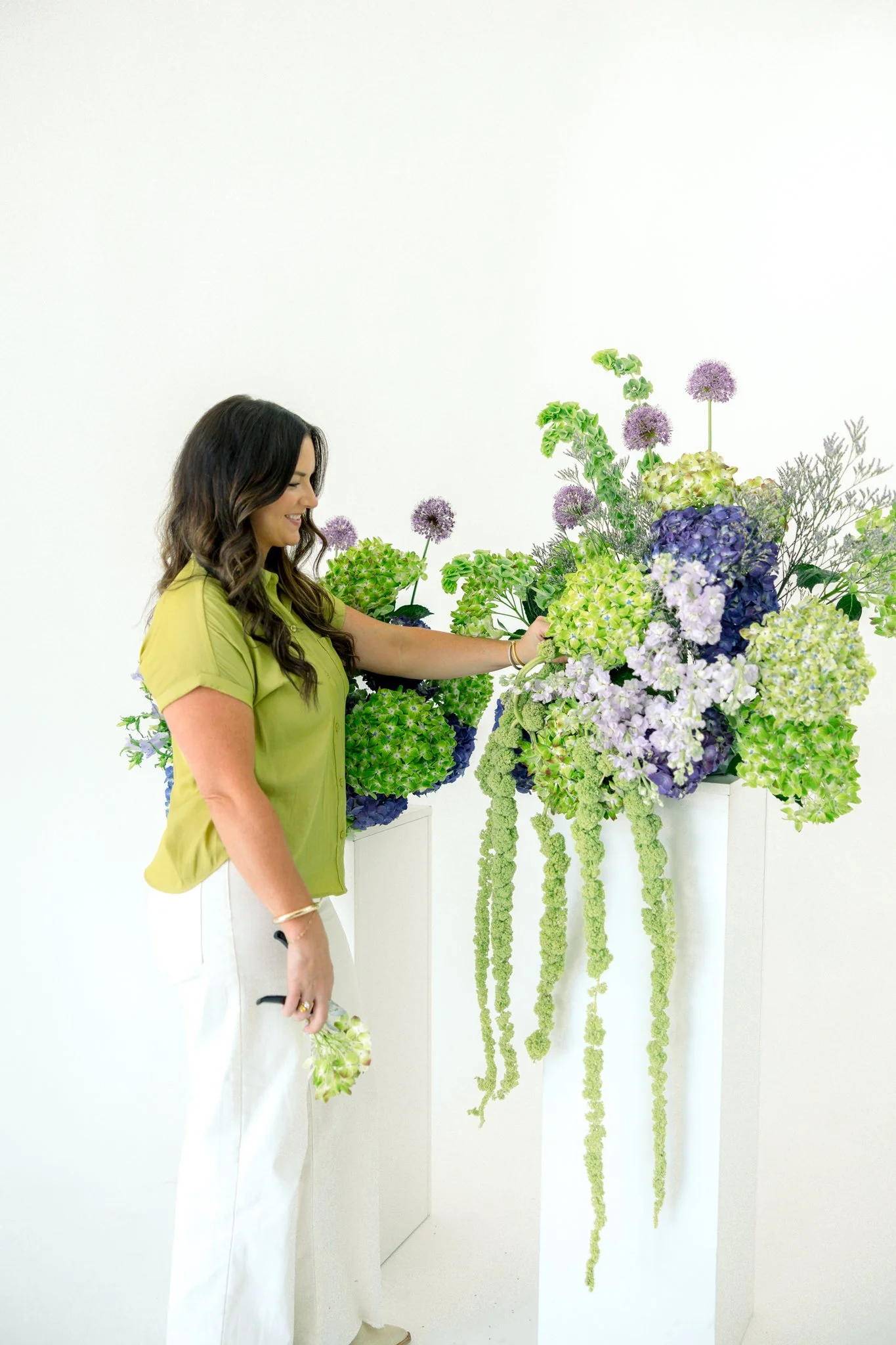 A woman with long brown wavy hair, wearing a green shirt and white pants, arranging a large floral art display with purple, green, and white flowers against a white background.