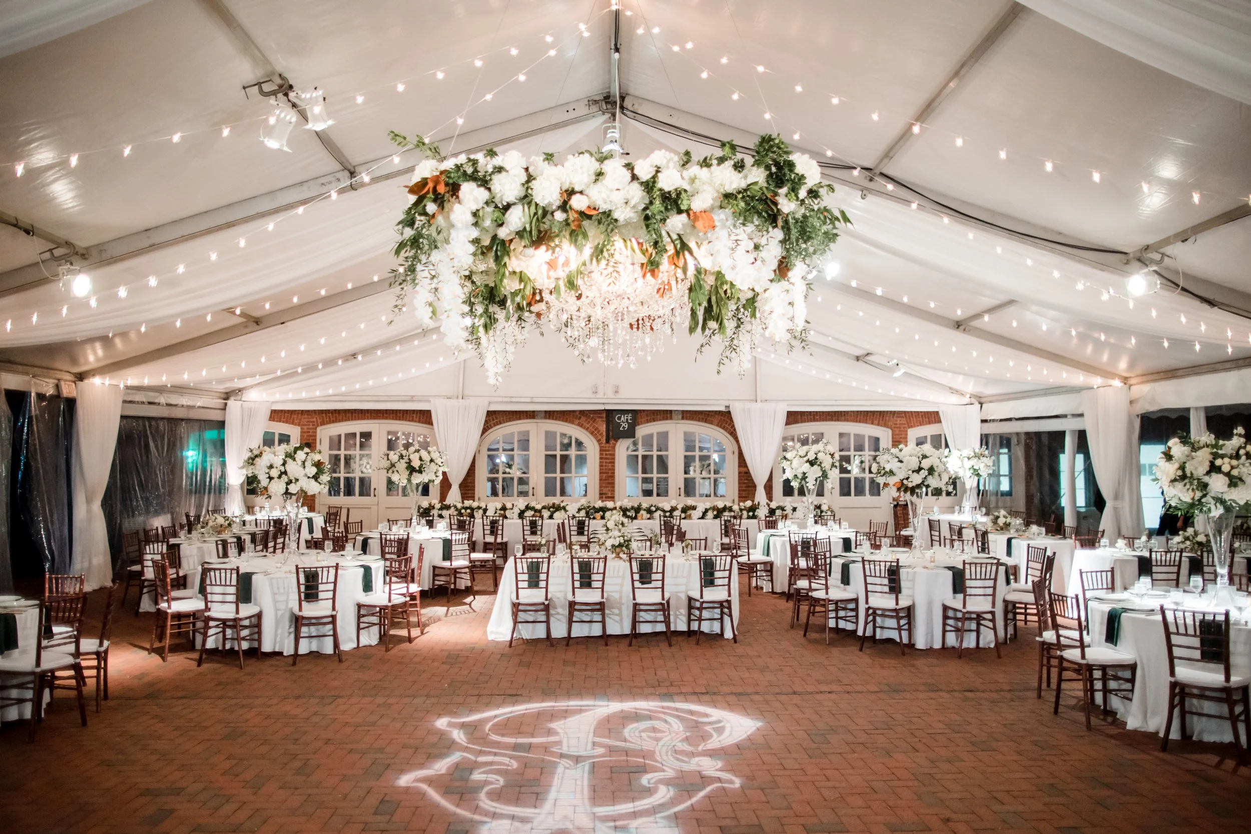 Elegant indoor wedding reception with tables, white tablecloths, brown chairs, and a floral chandelier hanging from the ceiling decorated with string lights.