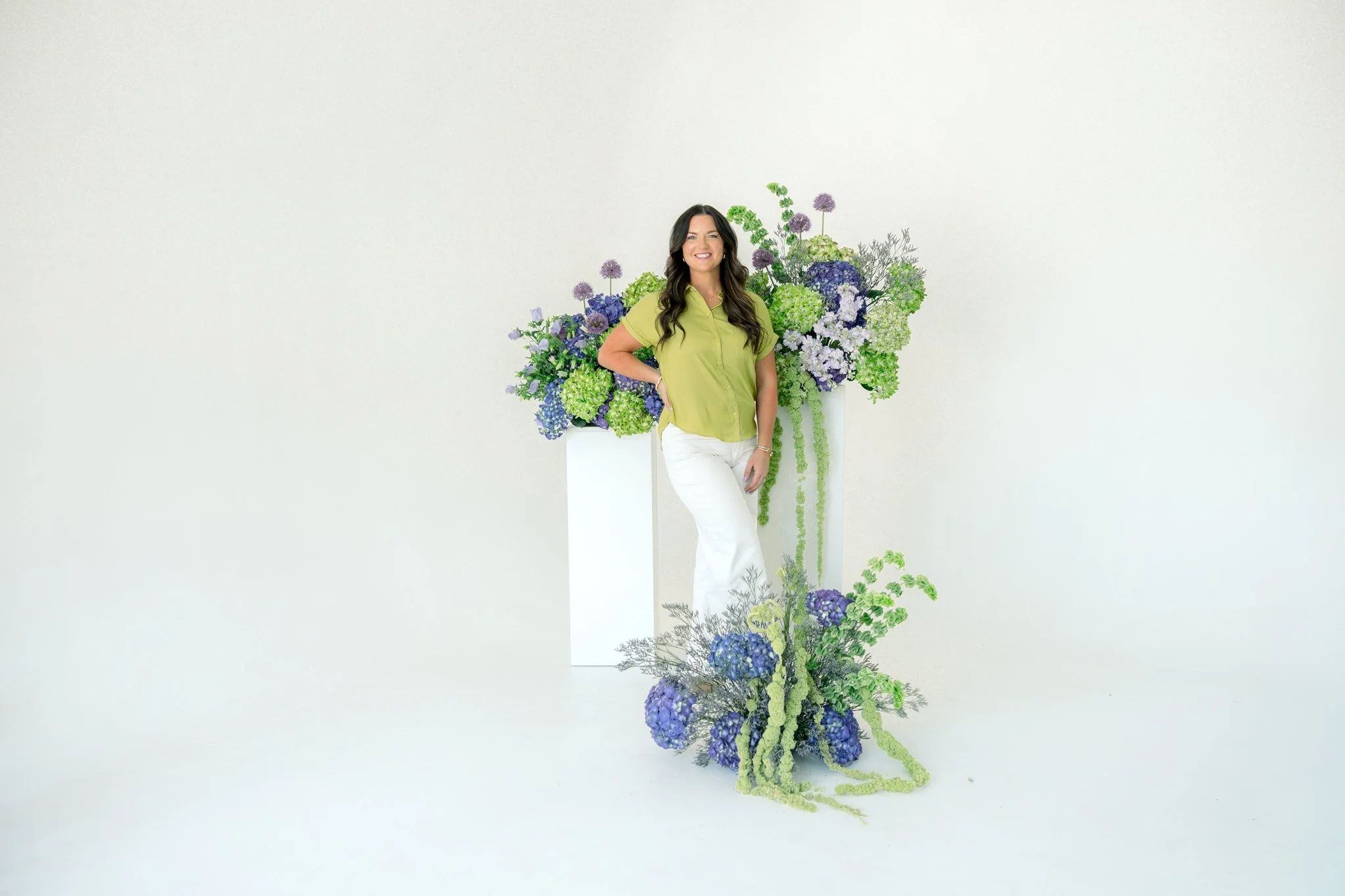 A woman in a lime green shirt and white pants standing beside a large, elaborate floral arrangement featuring various shades of purple, green, and blue flowers against a plain white wall.