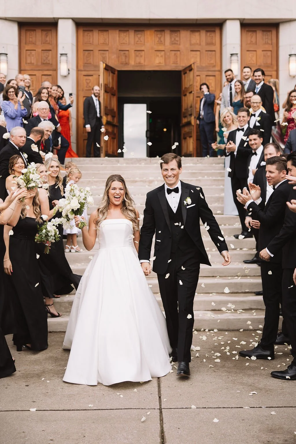 Happy newlywed couple walking down the steps of a church, surrounded by friends and family throwing flower petals.