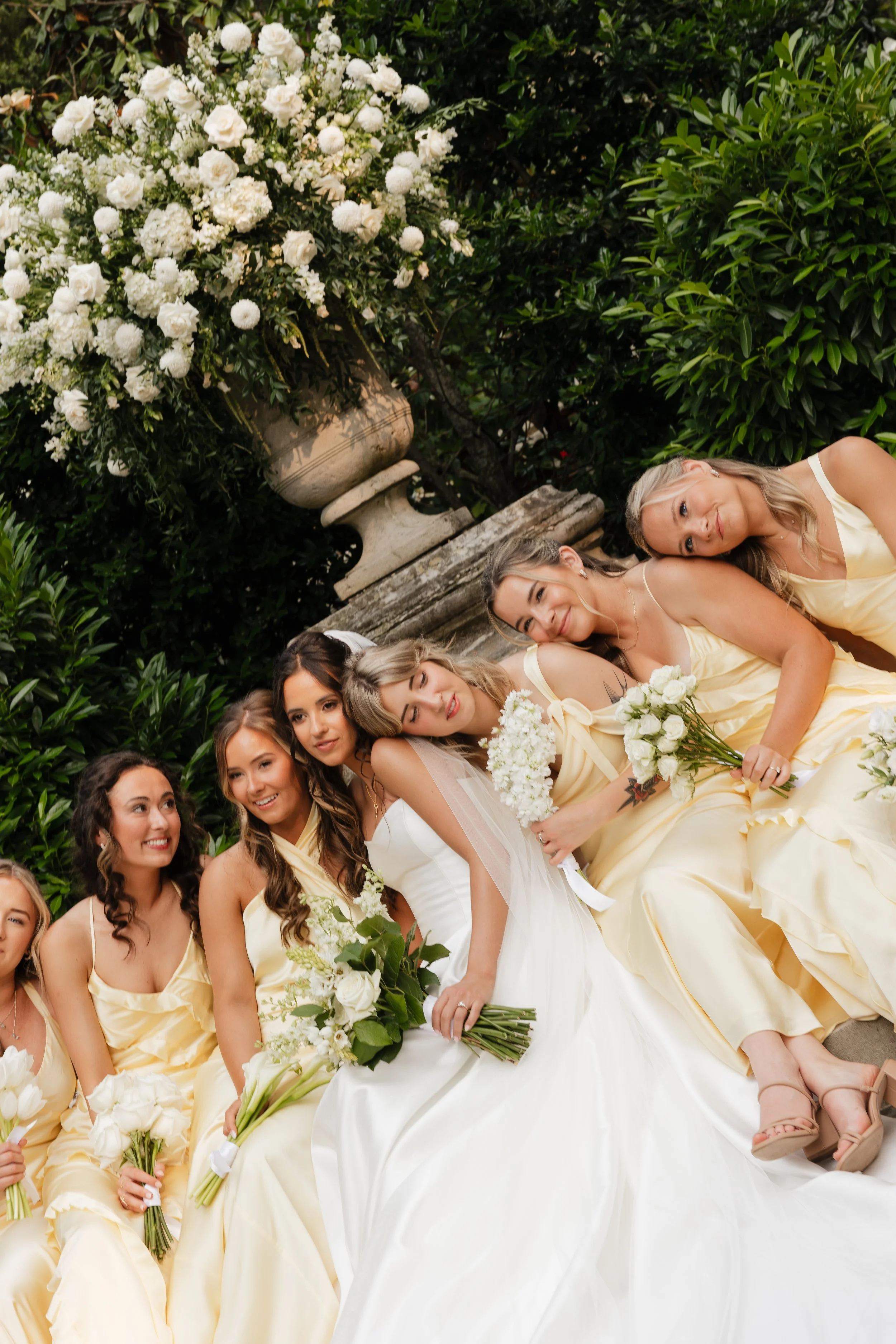 Group of women in yellow dresses and a bride in a white wedding gown with veil, holding white bouquets, sitting outdoors among greenery and flowers.