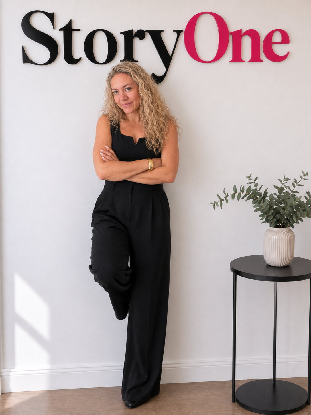 A woman with curly blonde hair wearing a black sleeveless top and black pants, standing with arms crossed in front of a white wall. Behind her, a sign reads 'StoryOne' with black and pink letters. To her right, there is a small black round table with a white vase holding green foliage.