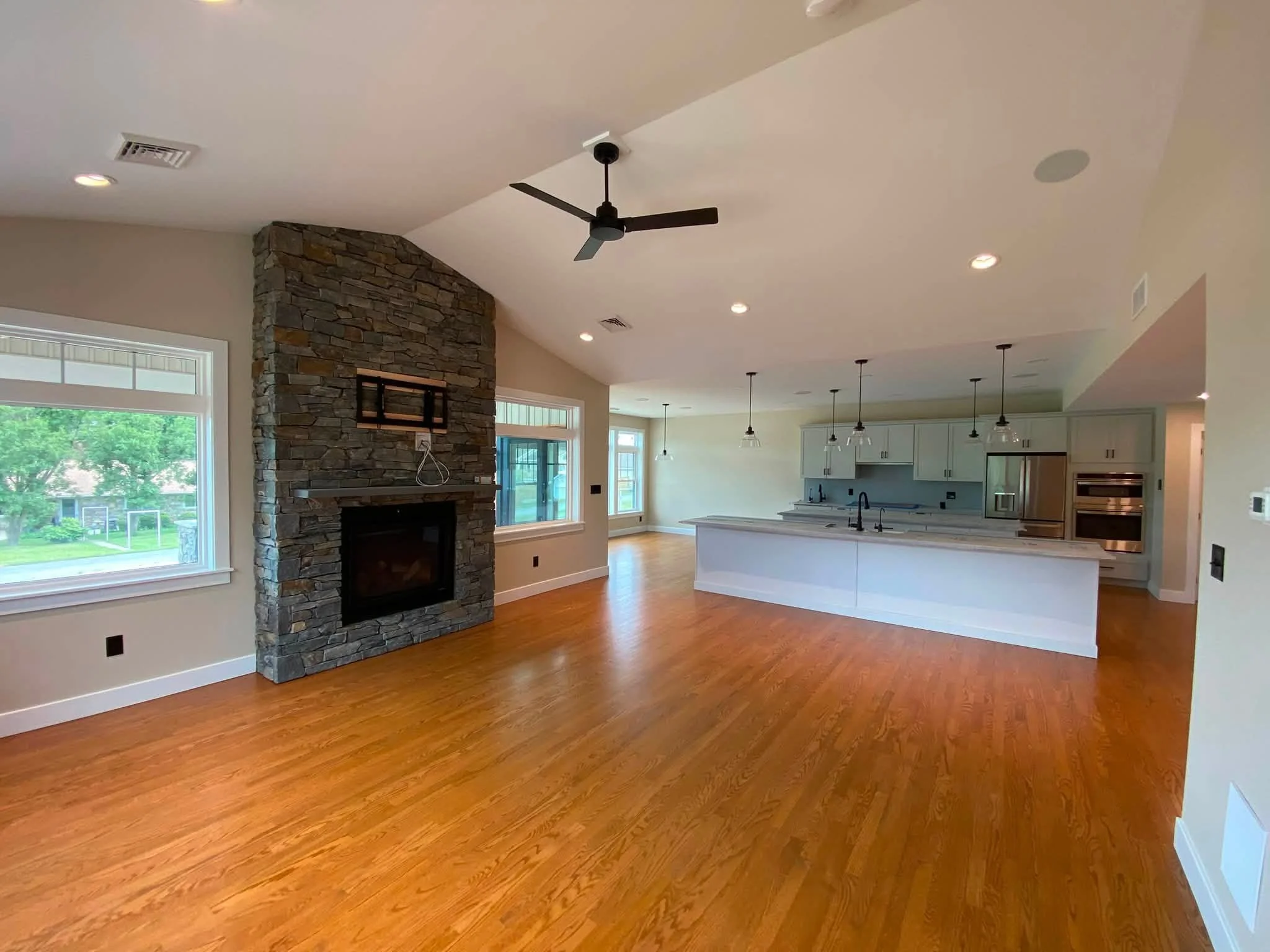 Open-concept living room and kitchen with hardwood floors, stone fireplace, ceiling fan, large windows, and hanging pendant lights.