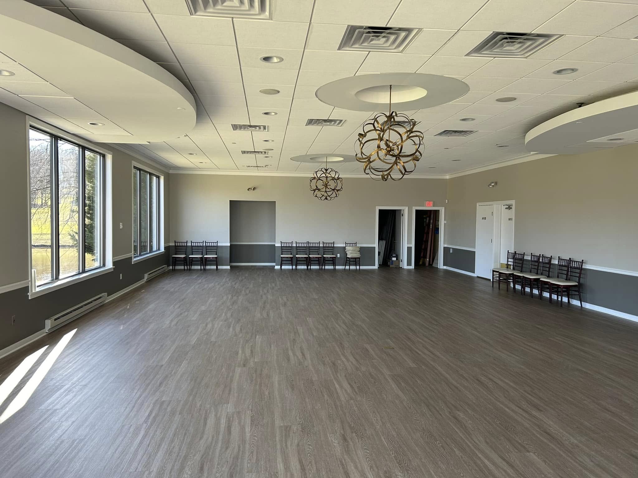 An empty banquet or event room with large windows, wooden flooring, beige walls, and modern circular chandeliers hanging from the ceiling.