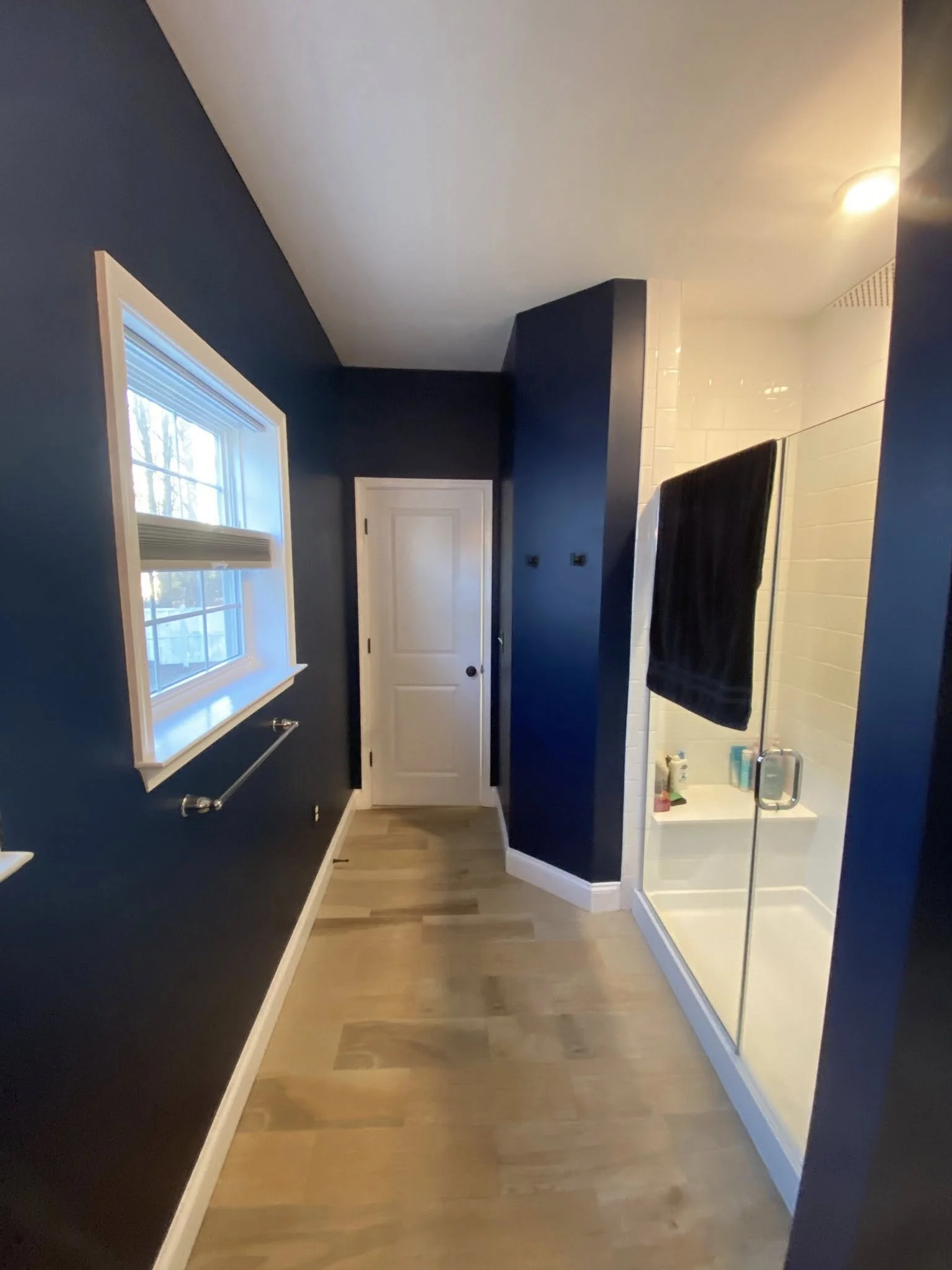 A narrow hallway with wooden flooring, dark blue walls, a white door at the end, a window on the left wall with blinds, and a bathroom with a glass door on the right side featuring a towel hanging inside and toiletries on the countertop.